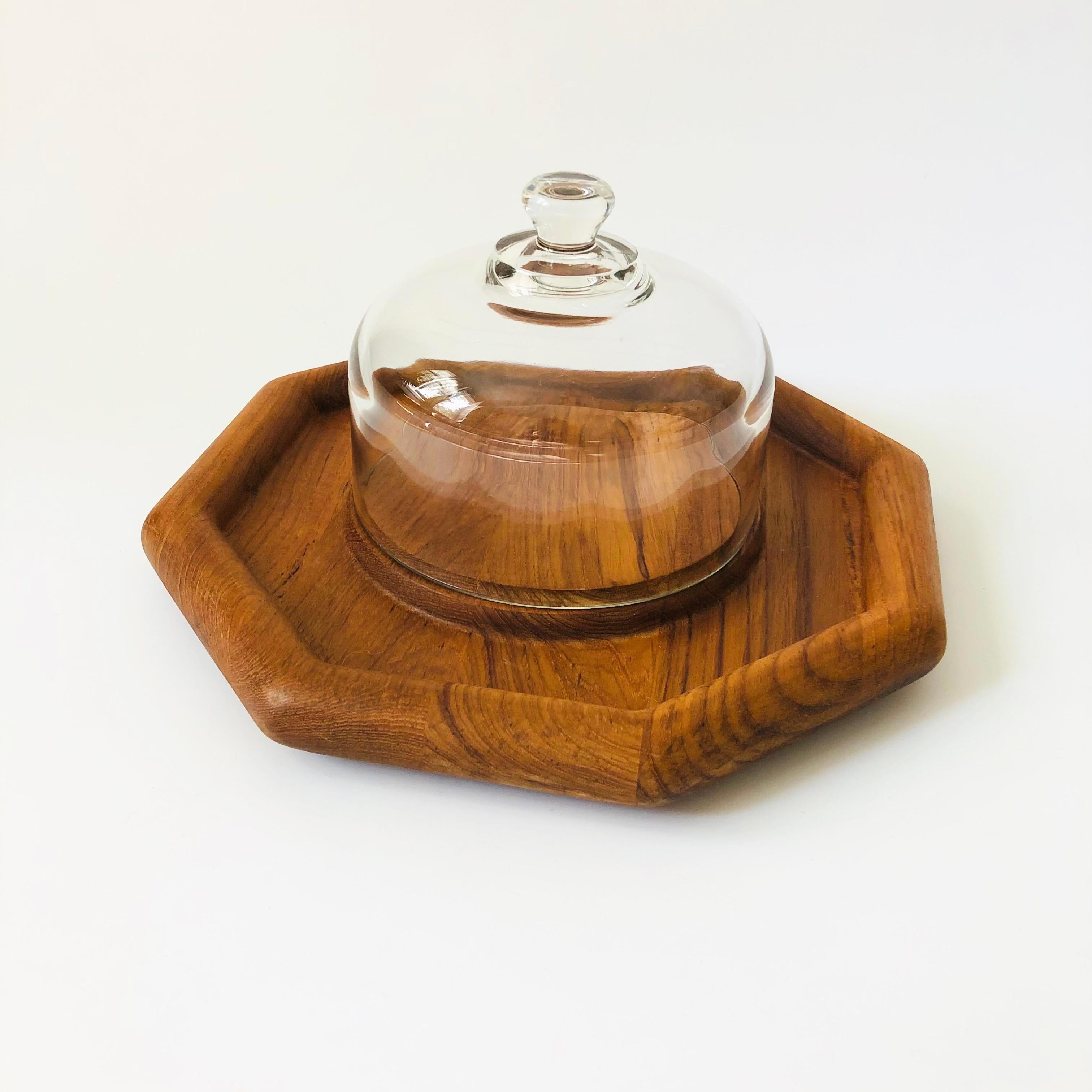 A vintage glass cloche on a teak tray. The top is made of heavy glass with an integrally formed handle sitting on an octagonal teak tray.  A useful serving piece perfect for serving appetizers or cheeses.

