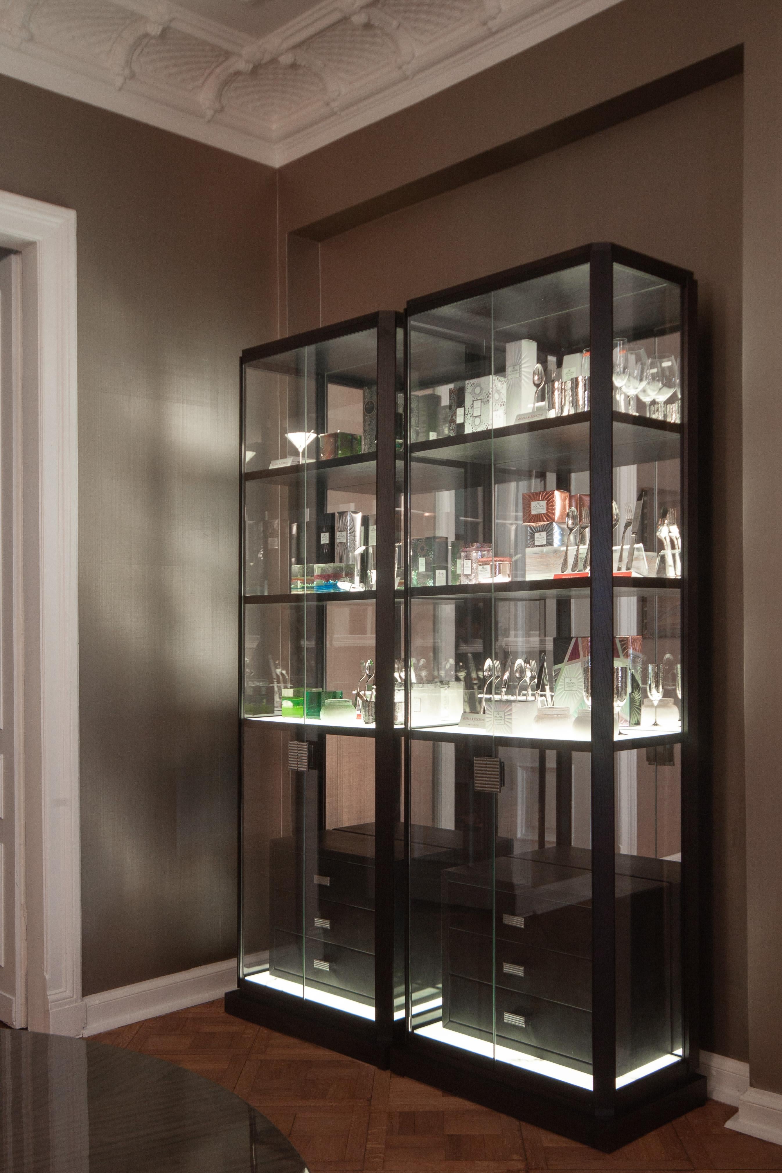 This glass cabinet's wooden elements are finished with a tinted walnut veneer. The shelves are made of translucent alabaster imitation material with lights embedded below the surface. The glass walls are around 3 sides, with a mirror in the