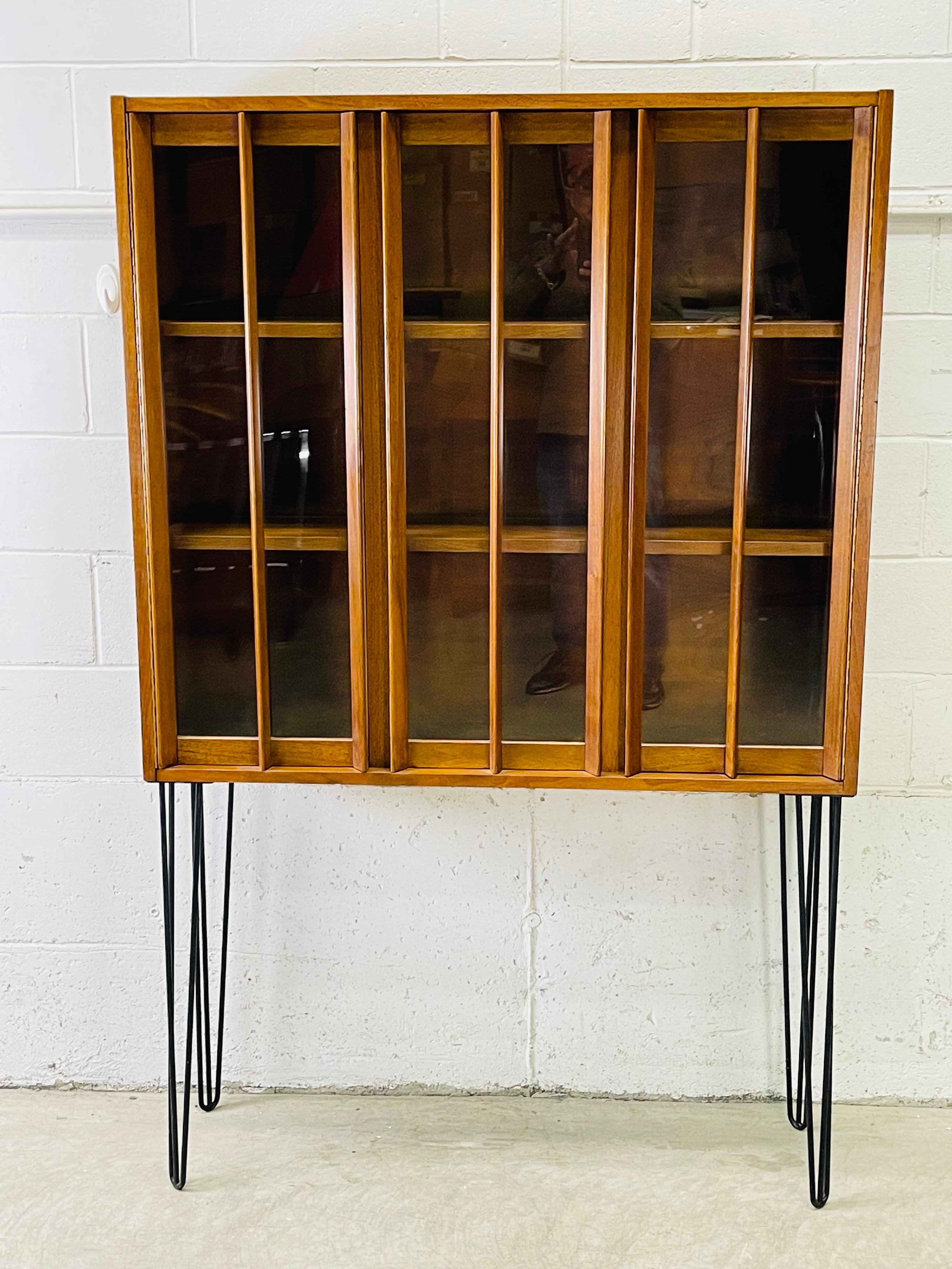 Vintage 1960s walnut wood and glass front tall display cabinet on iron hairpin legs. There are three sections for display and the shelves are 10” deep. The shelves are not adjustable. No marks.