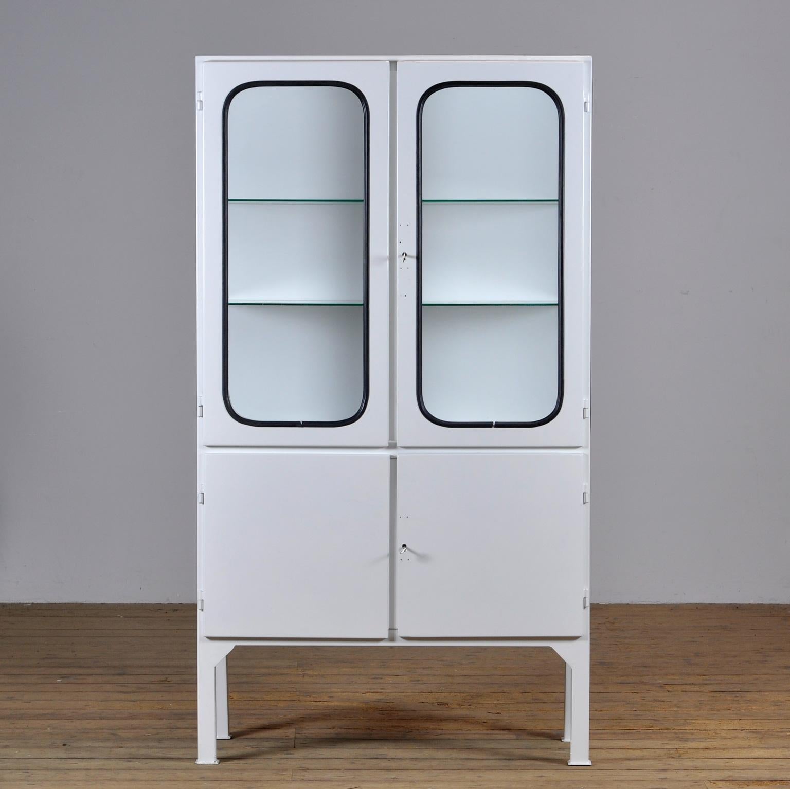 This medical cabinet was designed in the 1970s and was produced circa 1975 in hungary. It is made from iron and glass, and the glass is held by a black rubber strip. The cabinet features two adjustable glass shelves and functioning locks. 
The