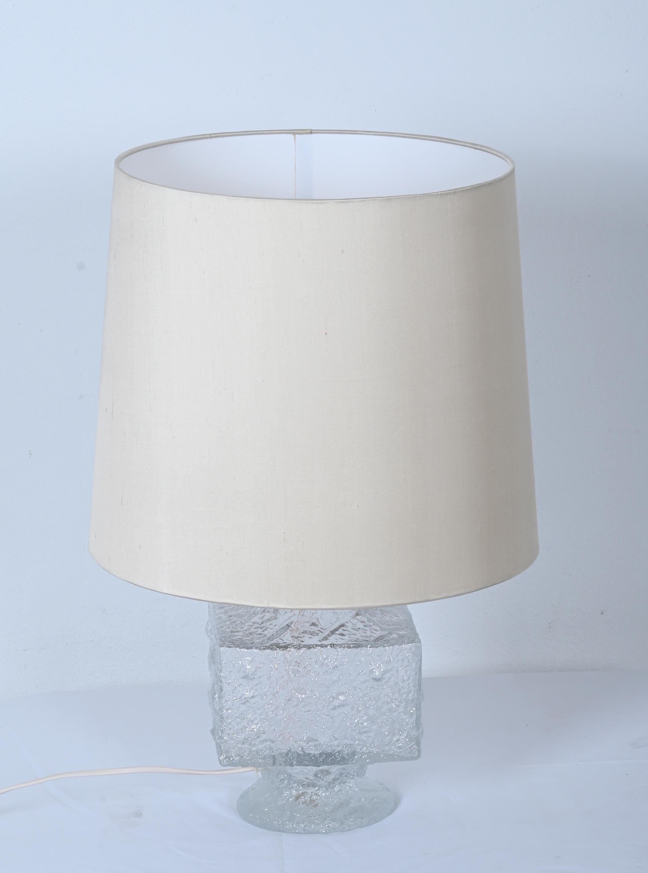 Base en verre moulé à colonne carrée décorée de cercles et de fossettes en bas-relief, équipée d'une prise E27 et d'un abat-jour en soie crème. Conçu par Timo Sarpeneva dans les années 1960 pour Lilux, Suède. 
