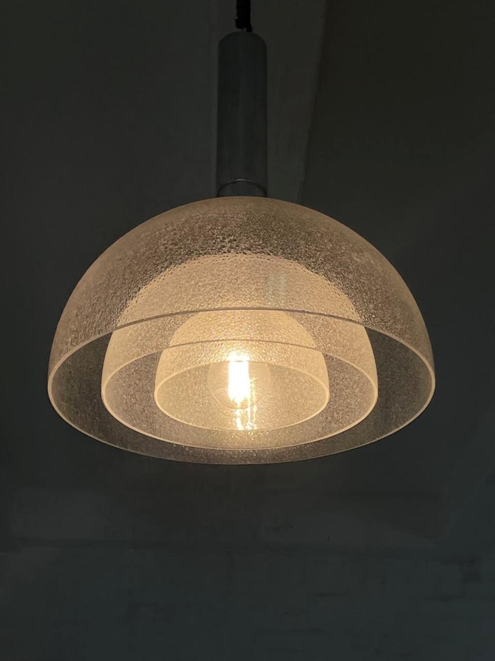Questa lampada a sospensione è stata progettata da Carlo Nason per l'azienda italiana di illuminazione Mazzega negli anni Settanta. Presenta un caratteristico paralume in vetro testurizzato a forma di cupola con uno stelo cilindrico cromato,