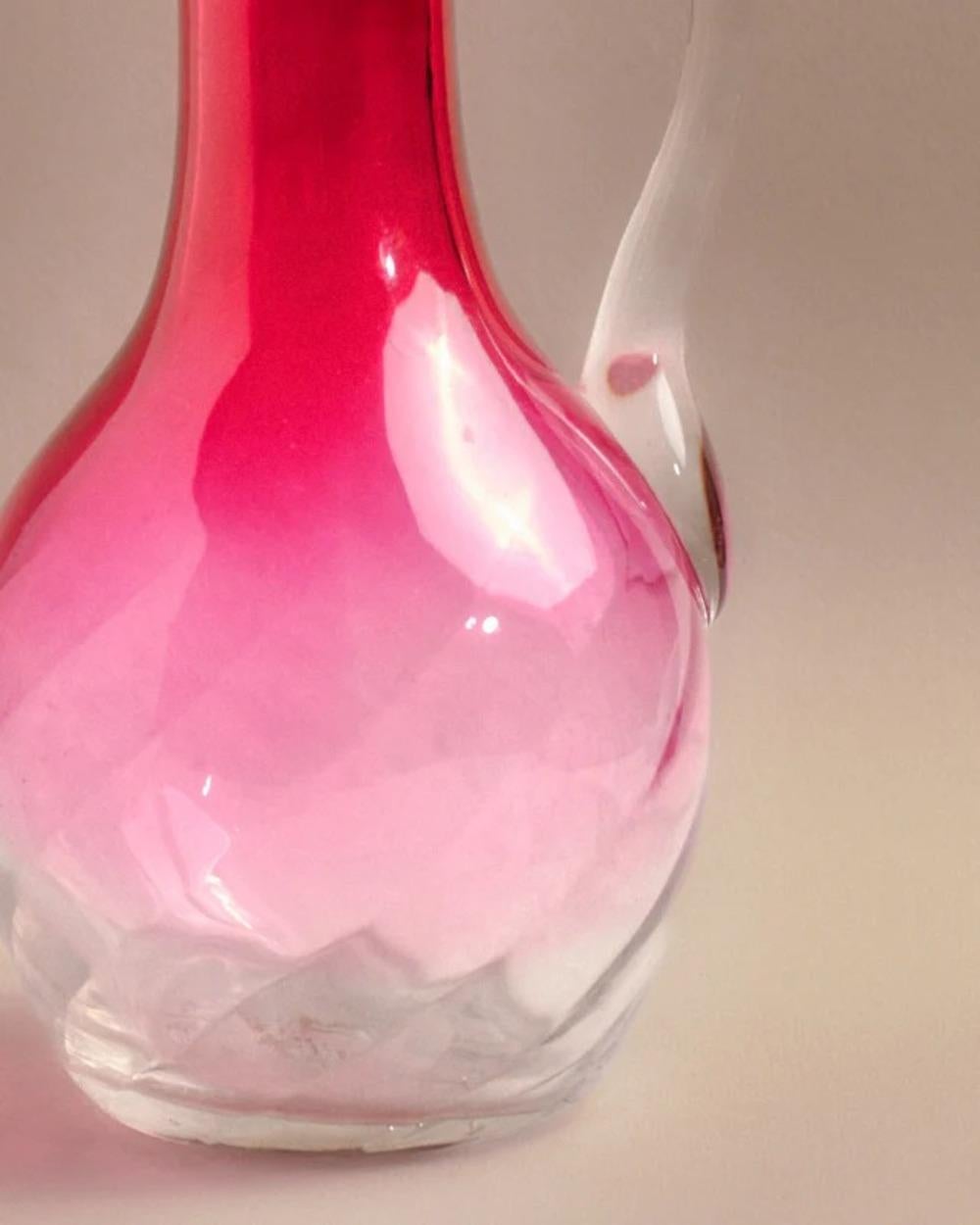 Krug aus geblasenem, rosa gefärbtem Glas, hergestellt in den 1970er Jahren. Der Körper weist im unteren Bereich regelmäßige Facetten auf, die dem Material geometrische Reflexe verleihen.
