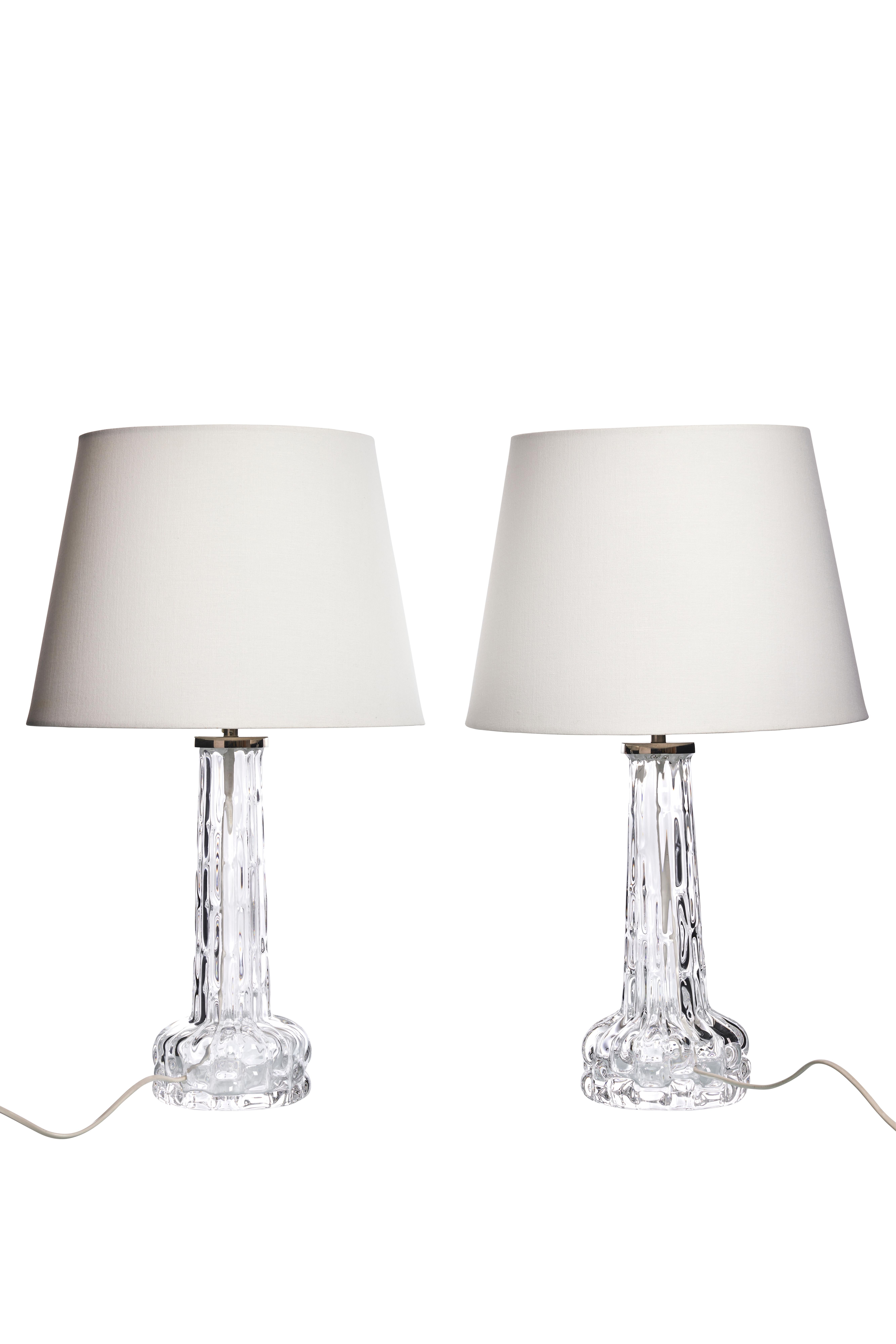 This set includes two vintage table lamps designed by Carl Fagerlund for Orrefors in the 1960s. Each lamp features a clear, textured glass base that adds a touch of Scandinavian elegance and craftsmanship to any space. 
The bases have a subtle
