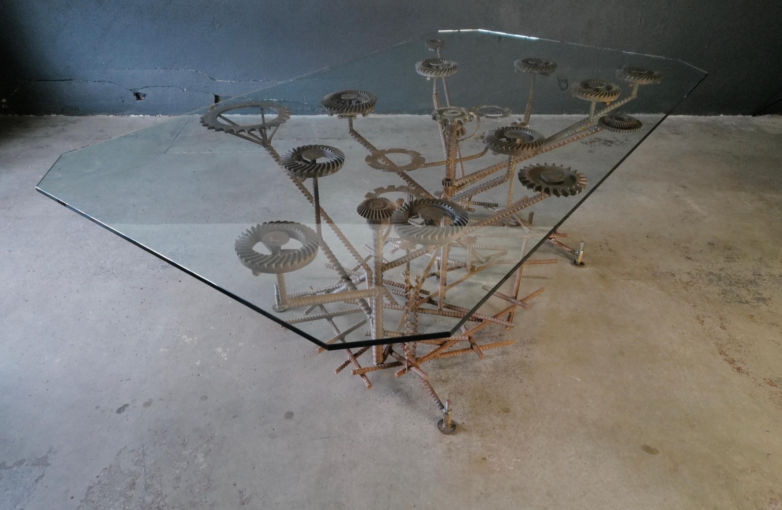 This stunning dining table features a spacious glass top that provides an elegant and modern surface for gatherings. Its unique base is a true work of art, crafted from a mesmerizing combination of rebar, gears, drill bits, and railroad nails. This