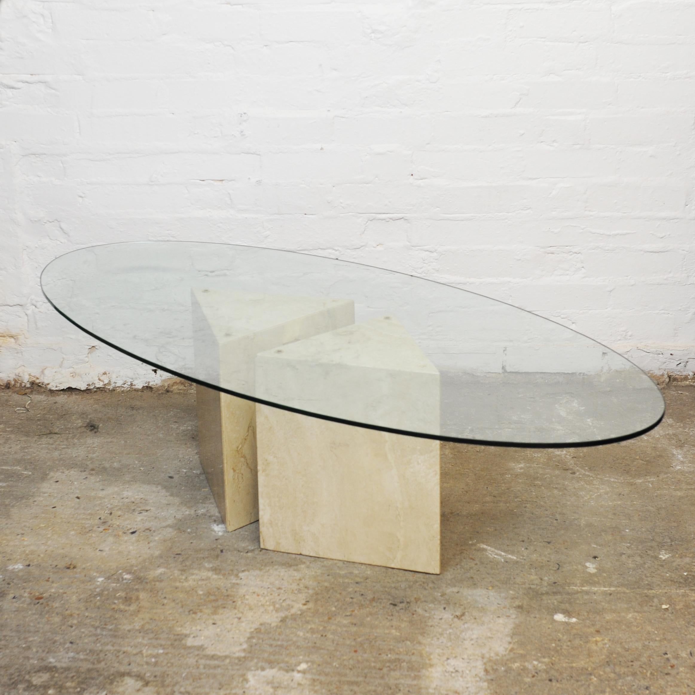 A post modern glass oval topped coffee table on two triangular bases joined by a brass piece. 

Designer - Unknown

Design Period - 1980 to 1989

Detailed Condition - Good with minimal defects. There is a chip on the travertine corner which is
