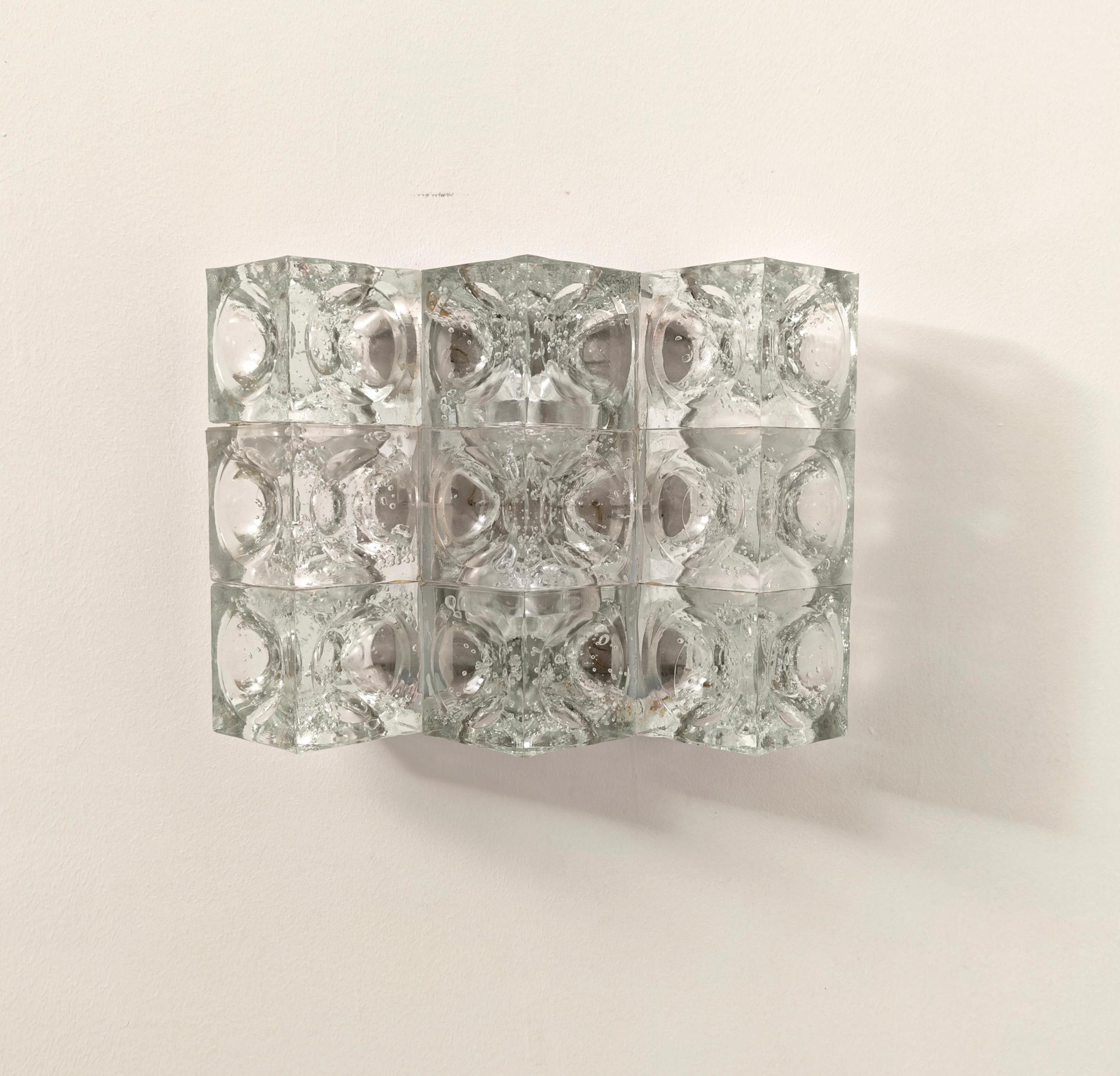 Questa applique, disegnata da Albano Poli per Poliarte, è composta da nove cubi di cristallo, ciascuno decorato con una superficie concava arrotondata. Le bolle d'aria sono sospese all'interno del vetro, creando una straordinaria diffusione della