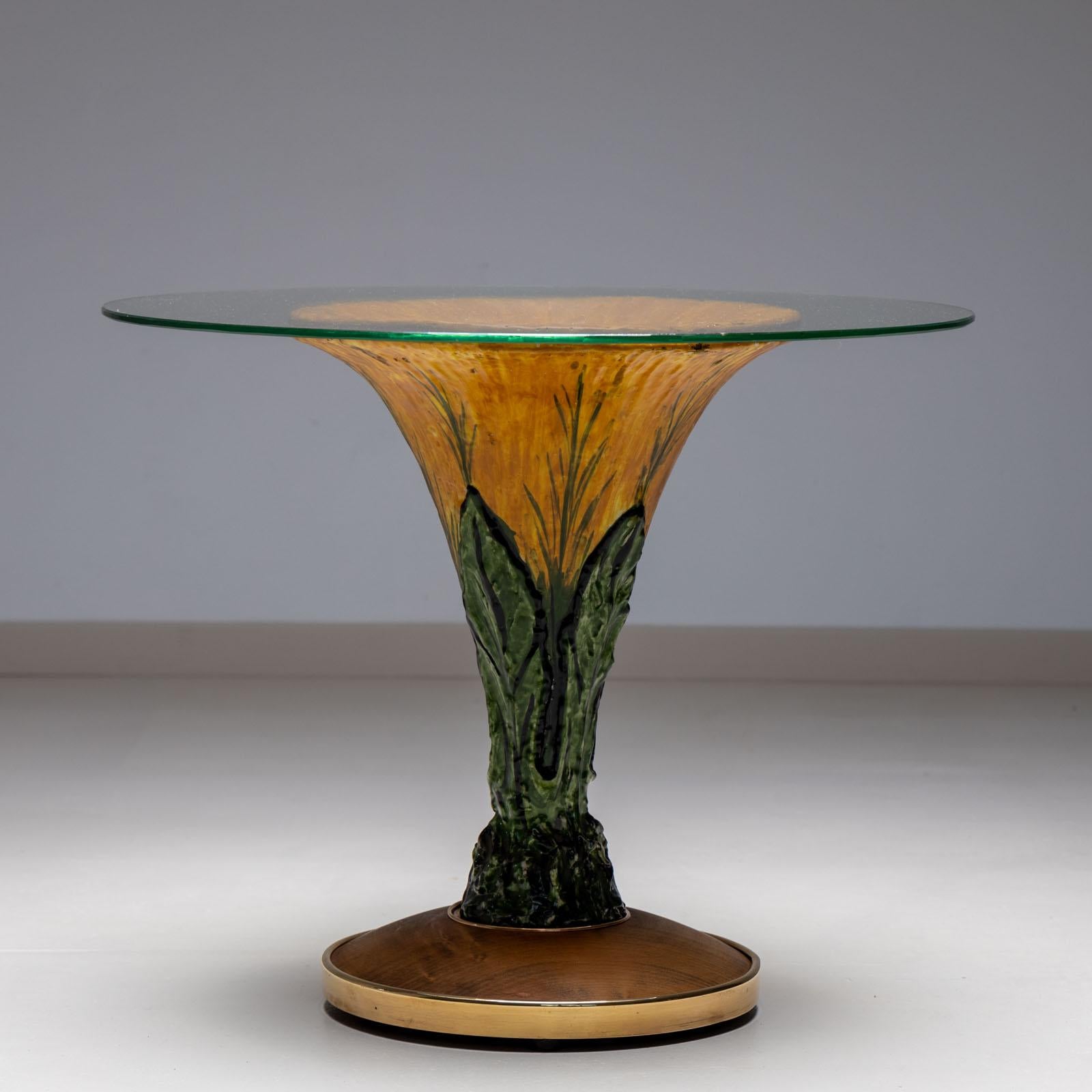 Tavolino italiano in ceramica degli anni '60, con piano rotondo in vetro. Il tavolo è realizzato in ceramica smaltata giallo-verde con motivi di foglie stilizzate in rilievo intorno al fusto a forma di tromba. Il tavolo poggia su un disco di legno