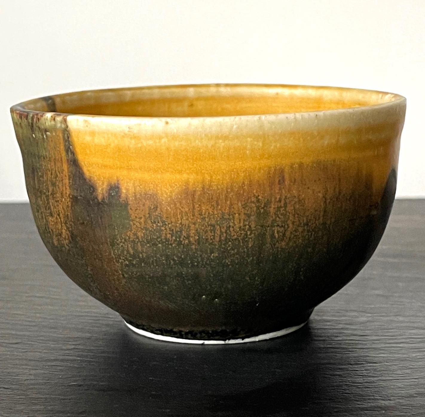 A ceramic tea bowl (chawan) by Japanese American artist Toshiko Takaezu (American, 1922 - 2011). In a classic chawan form, this small tea bowl strikes the viewer with its beautifully applied glaze. On its golden-ochre background, an ink like black