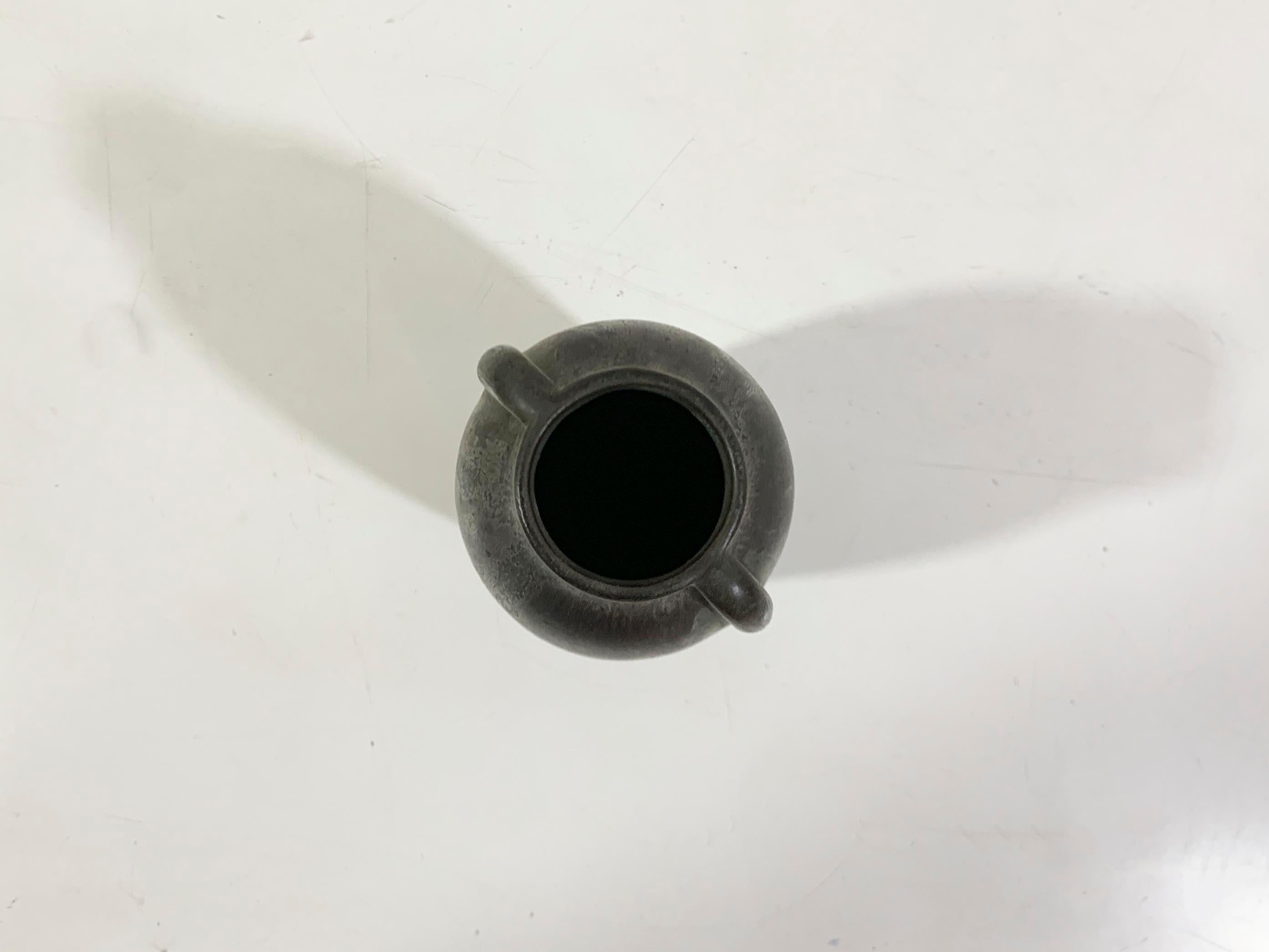 Questo vaso è un pezzo di ceramica lavorato a mano con uno smalto scuro e una finitura organica leggermente strutturata. La sua forma elegante e allungata è accentuata da due piccoli manici a cappio vicino al collo, che le conferiscono un design