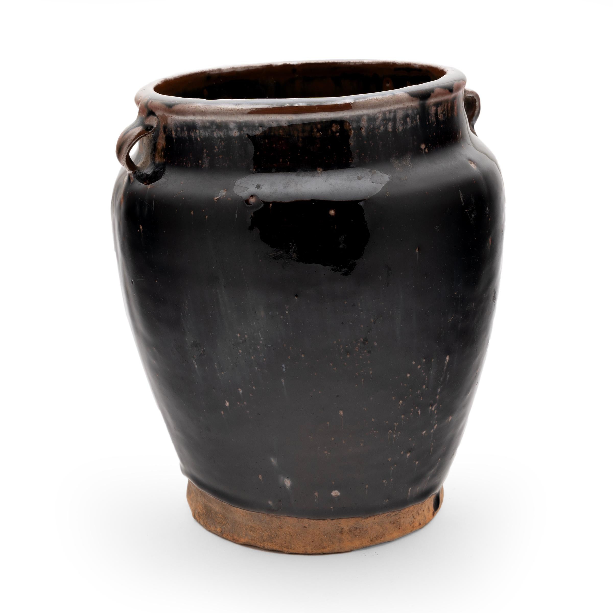 A glossy black glaze clings to the form of this slender kitchen jar's banded surface. The glazed vessel dates to the early 20th-century and was used for storing food and condiments in a provincial Qing-dynasty kitchen, as evidenced by its interior