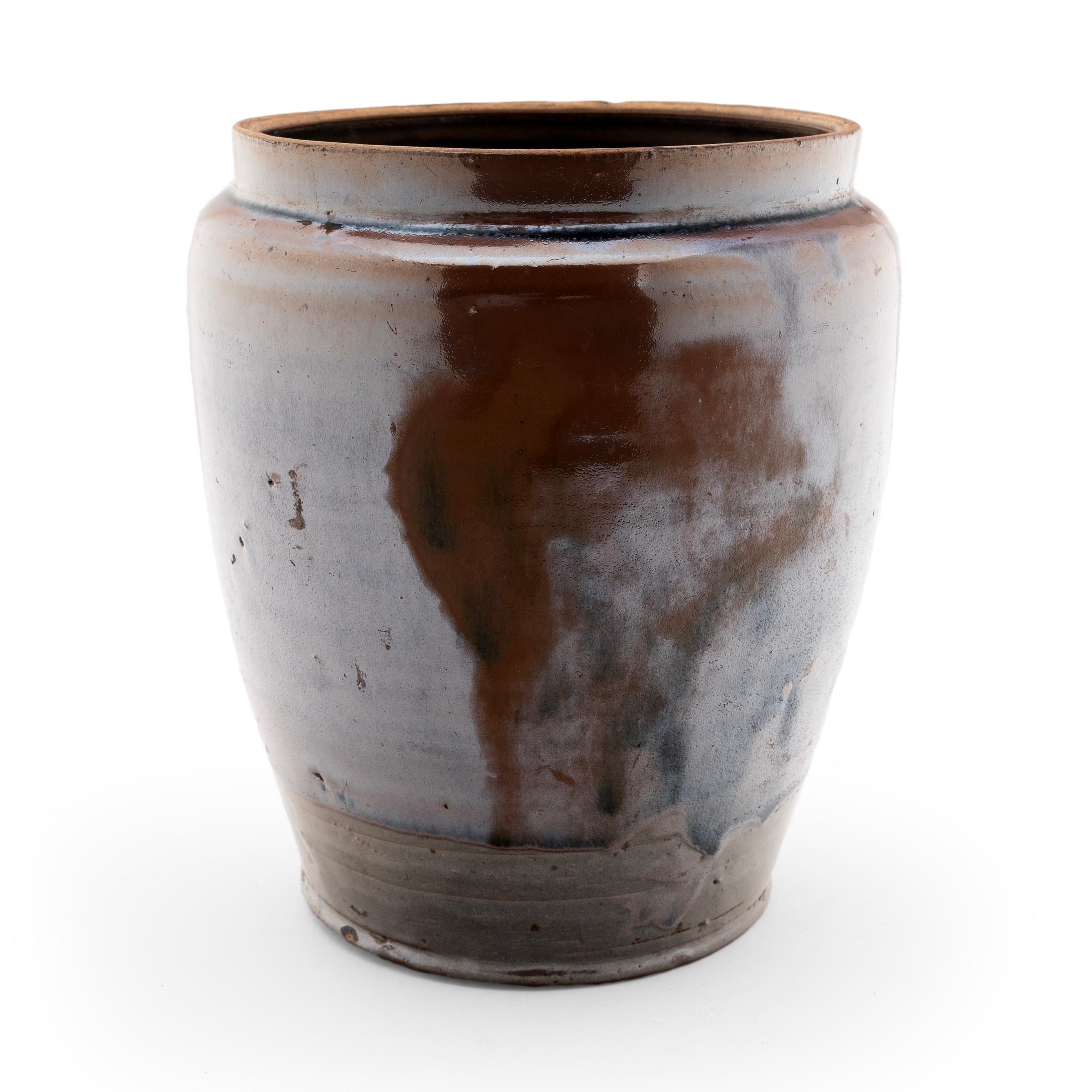 A thick, glossy brown glaze coats the tapered form of this early 20th-century terra cotta kitchen jar, lingering on imperfections and glistening with an iridescent sheen. As evidenced by the glazed interior, this wide-mouth jar was once used daily