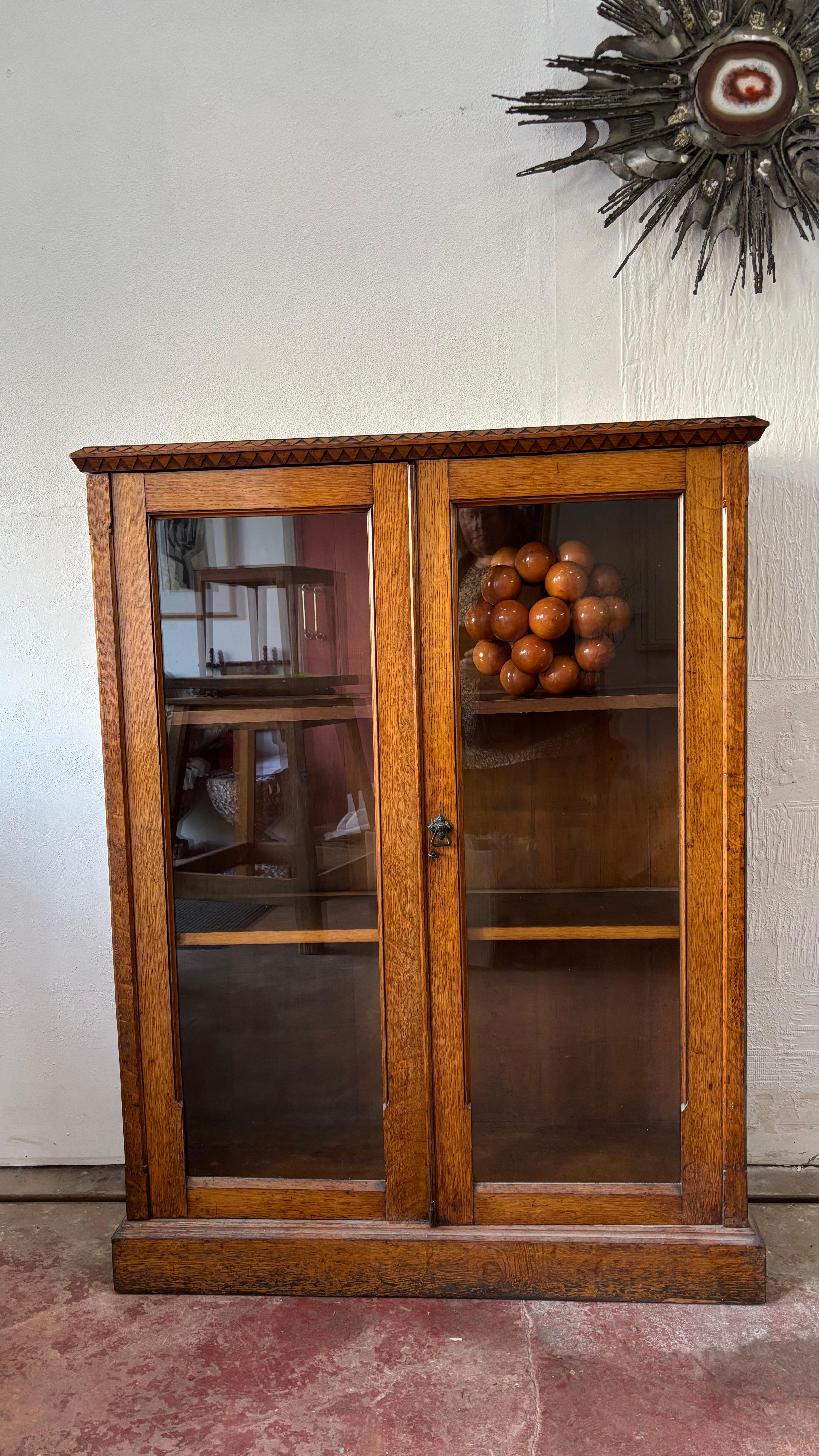 A really nice honey oak Arts and Crafts glazed front display cabinet. The top frieze has an attractive zig zag frieze. The two glazed doors feature the original Arts and Crafts ironwork handle, a lock and key. They open and inside there are two