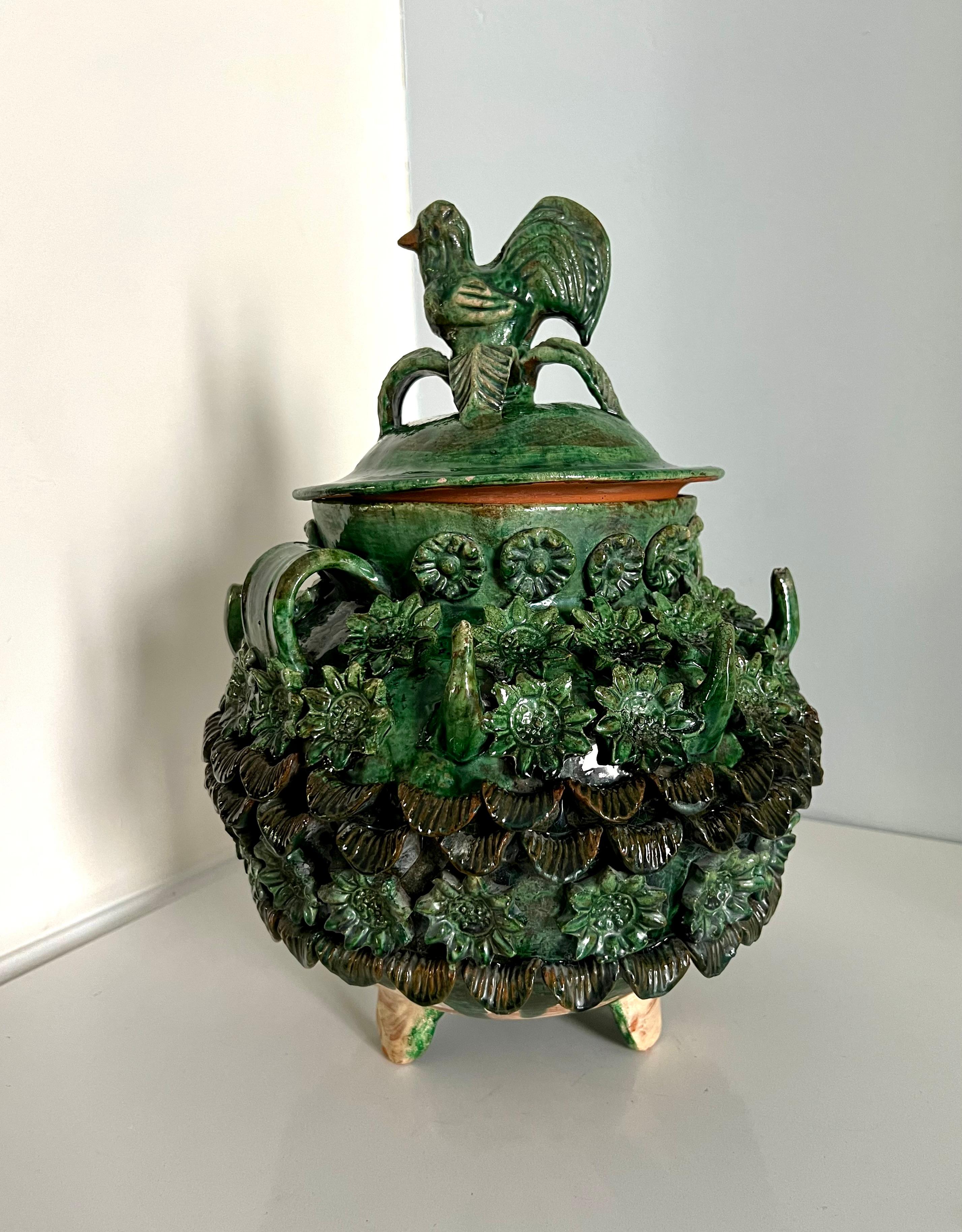 A unique Lidded terracotta majolica style jar with three dimensional flowers around and a rooster atop the lid. The container could be applicable but likely more decorative. The piece stands on three pod feet. A wonderfully attractive and decorative