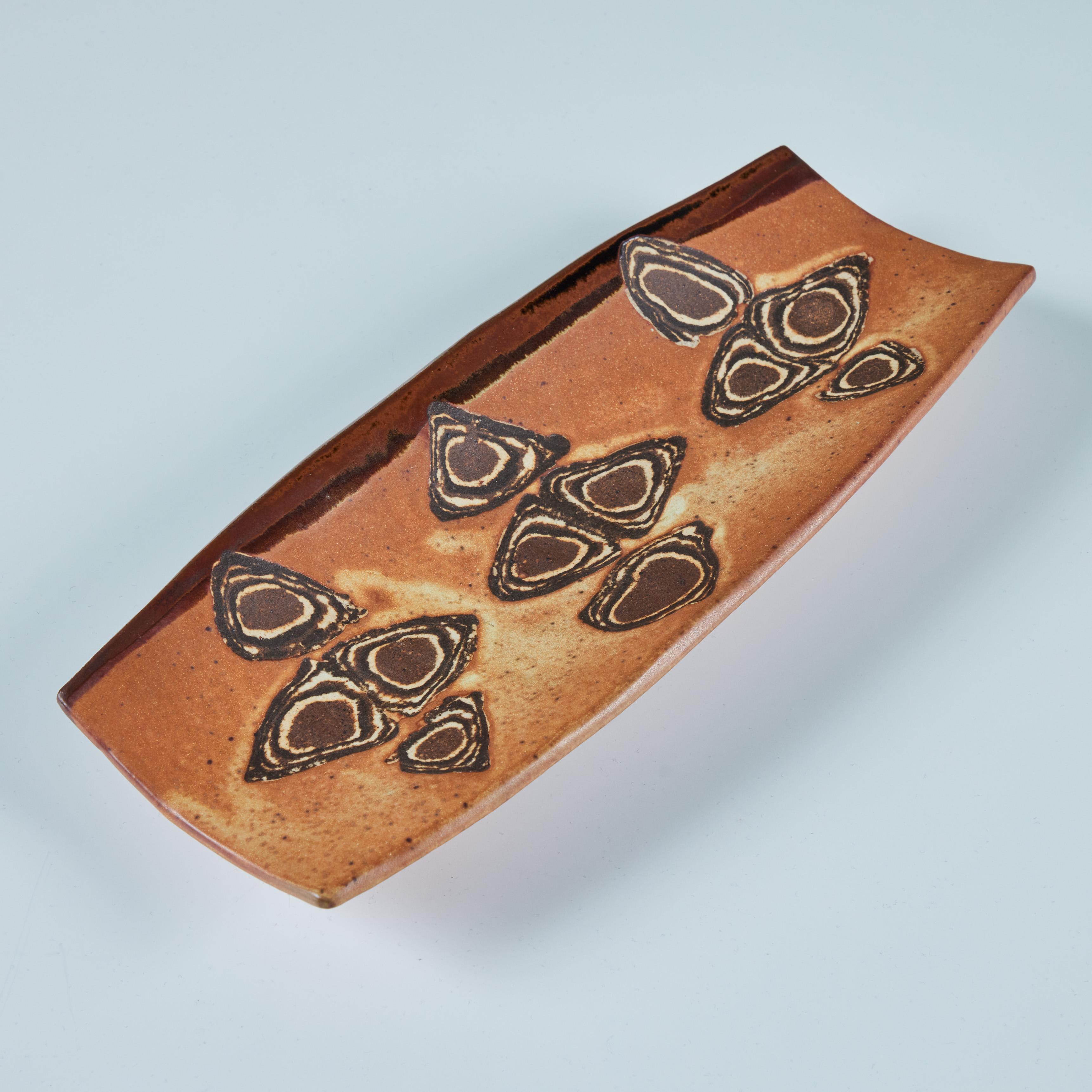 Stoneware plate features a rectangular shape with turned two curled sides. The plate is glazed in burnt orange and brown tones with speckling throughout. The piece has a three geometric design depicting triangles and diamonds. It rests on three