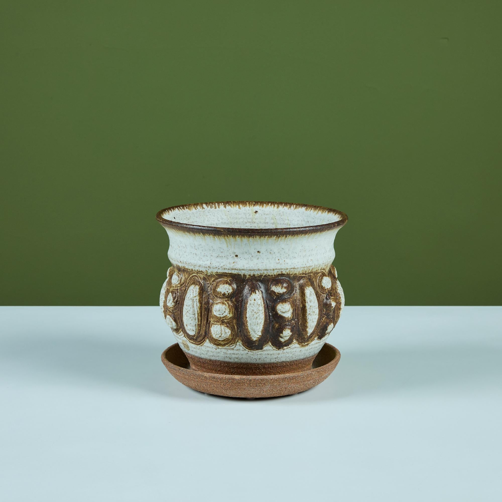 Recipiente in gres smaltato con presa d'acqua. Questo pezzo è caratterizzato da rientranze delle dita su tutto il corpo che creano un motivo geometrico di cerchi e ovali. È rifinito con uno smalto color crema e marrone, con annesso un piatto di