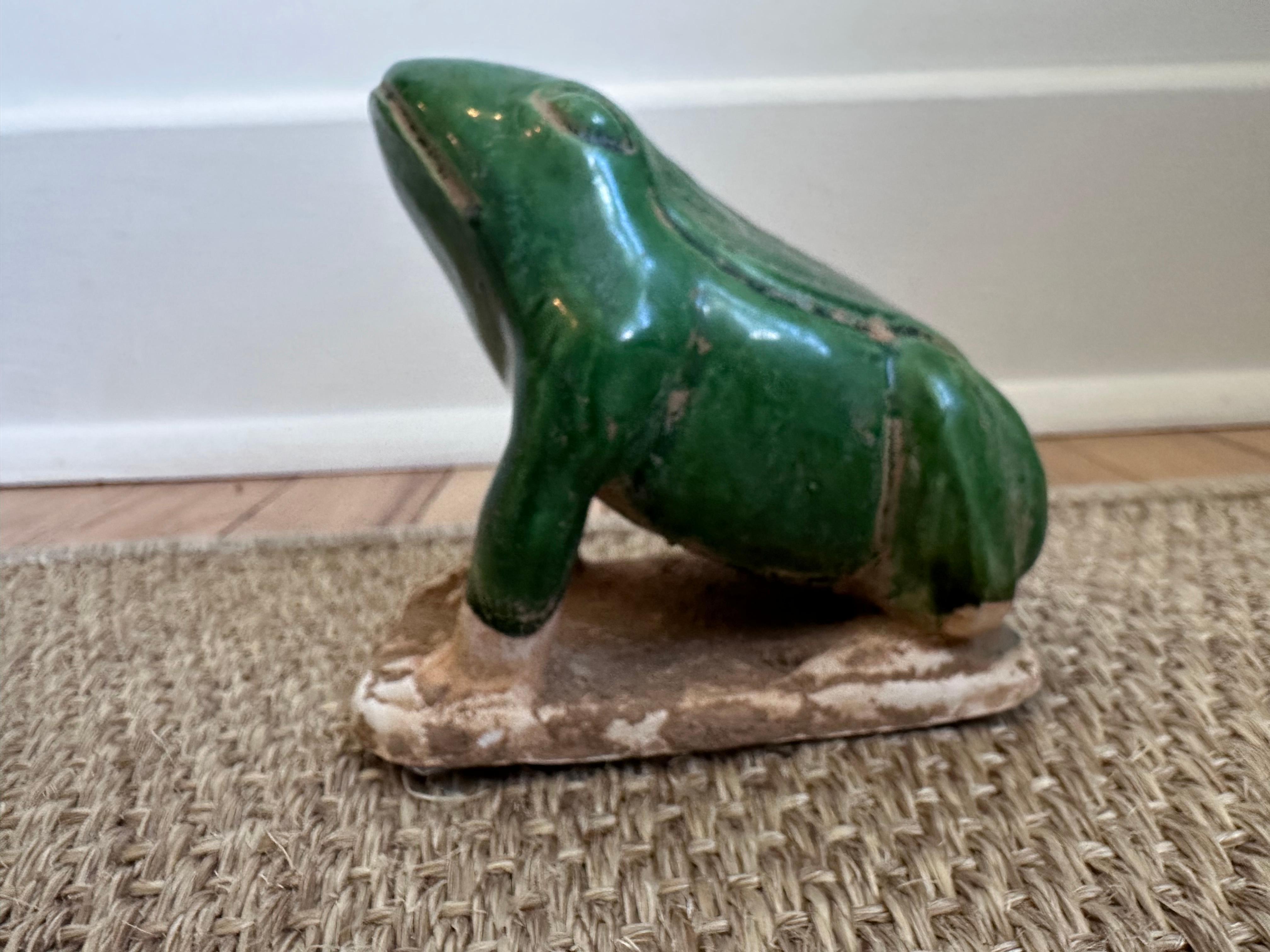 Voici une charmante statue de grenouille en céramique au caractère rustique et usé par le temps.

La grenouille est représentée en position assise ou accroupie, la tête légèrement inclinée vers le haut, ce qui lui confère un sentiment de vigilance