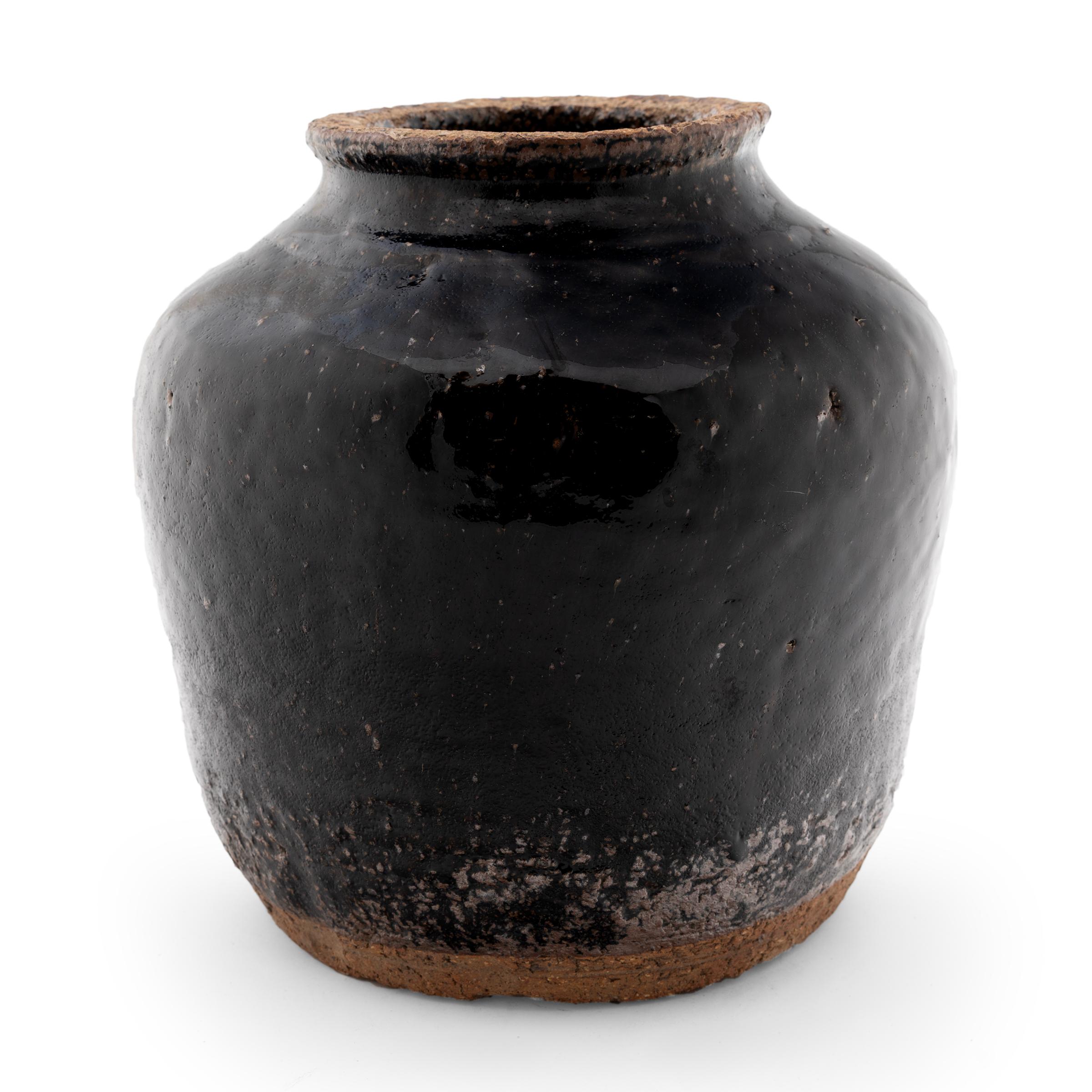 Crafted in the early 20th century, this glazed jar was once used in a Qing-dynasty kitchen to store vinegar, wine, or condiments, as evidenced by its narrow, constricted neck and interior glaze. The jar boasts a thick, dark black glaze over its high