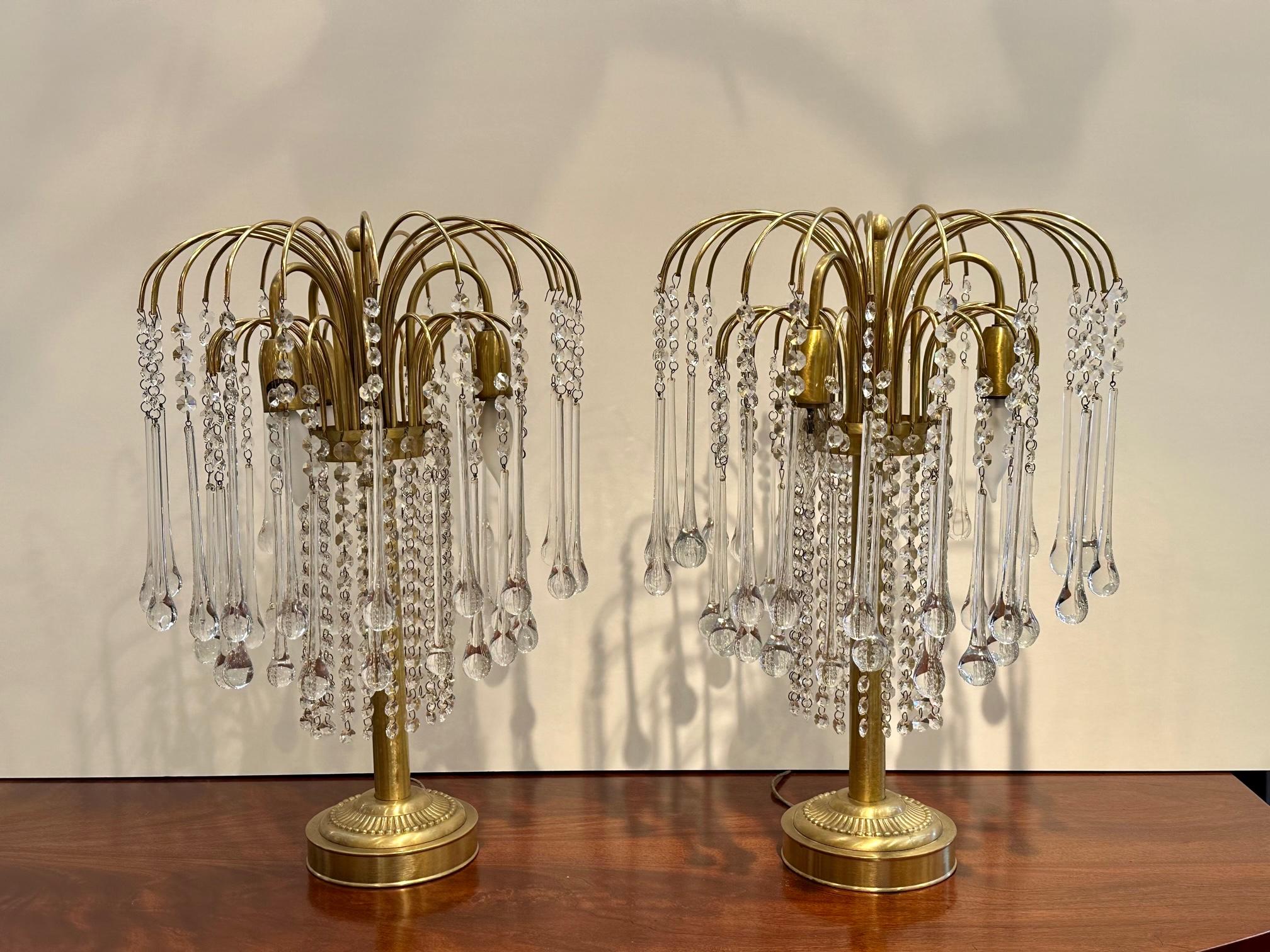 Fabulously glamorous decorative pair of Italian brass and crystal table lamps having an overflowing cascade of tear drop crystals erupting from arc shaped bands of brass. 3 lights per lamp.