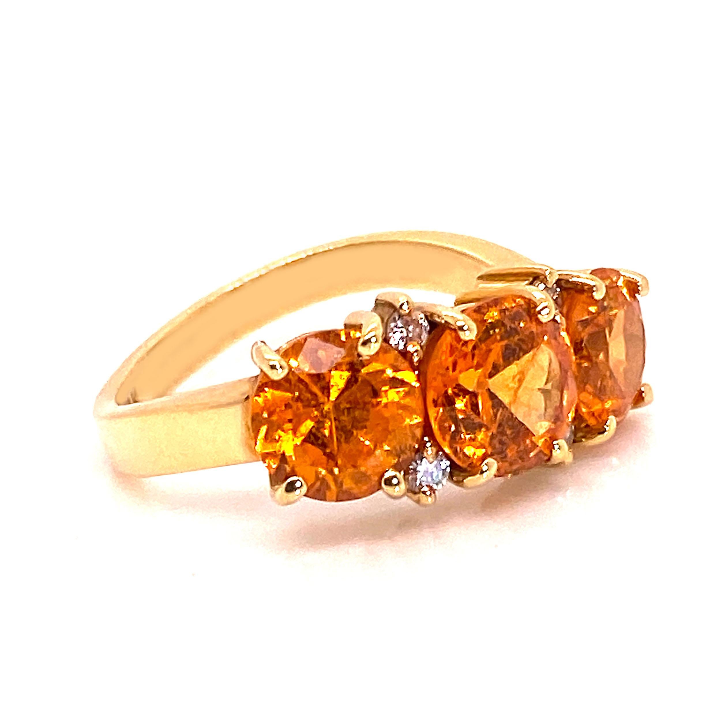 Hand made 18K yellow gold ring with three round 6.5-7.5 MM brilliant orange Spessartite garnets of approximately 4.5 carats total weight. The sparkling Spessartite garnets are enhanced by four diamonds of approximately 0.08 carats total weight. 