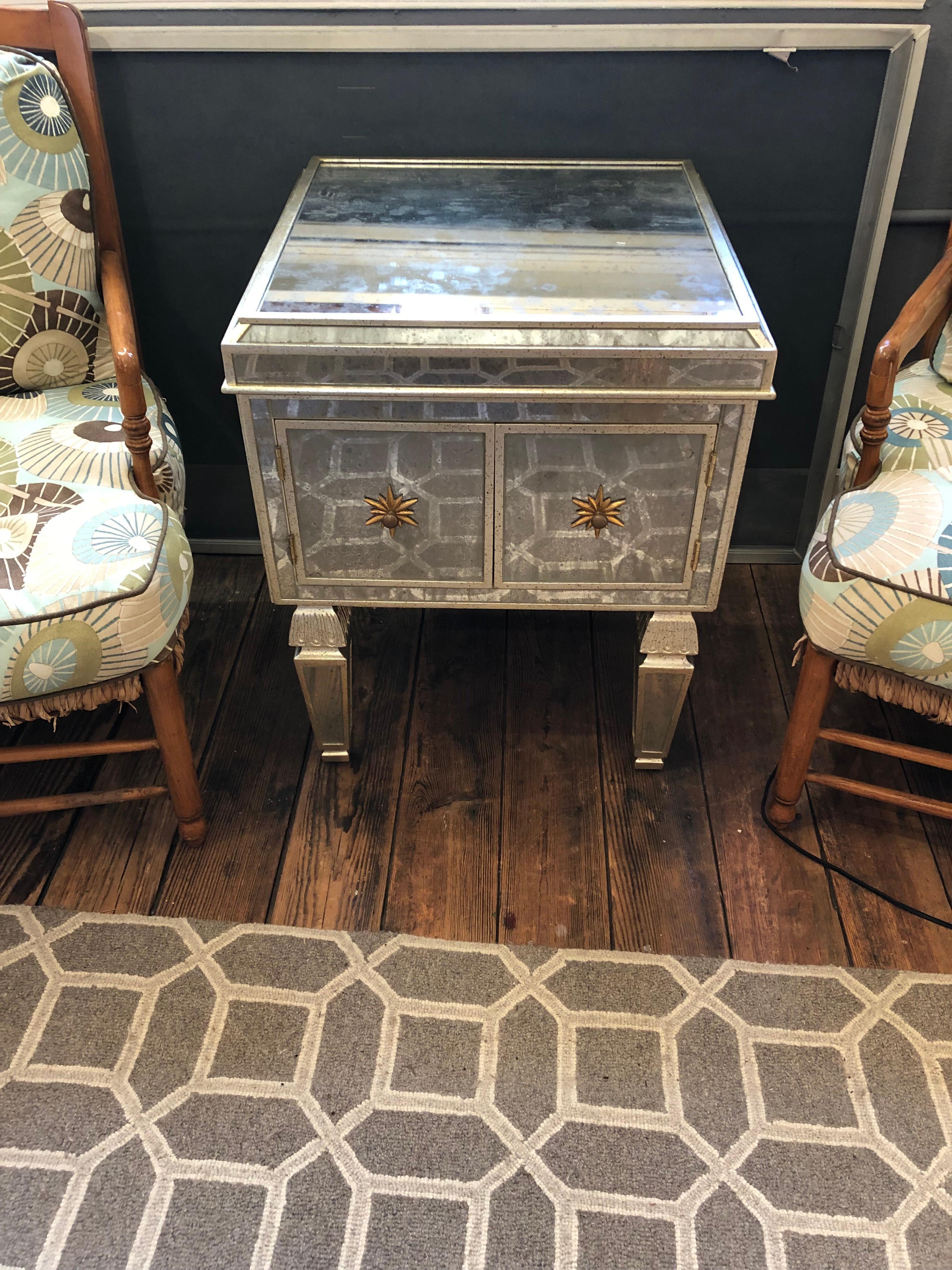 A glamorous square aged mirror side table cabinet having two doors that reveal storage inside, handsome mirrored sculpted legs, and contrasting sunburst gold knobs on the doors.