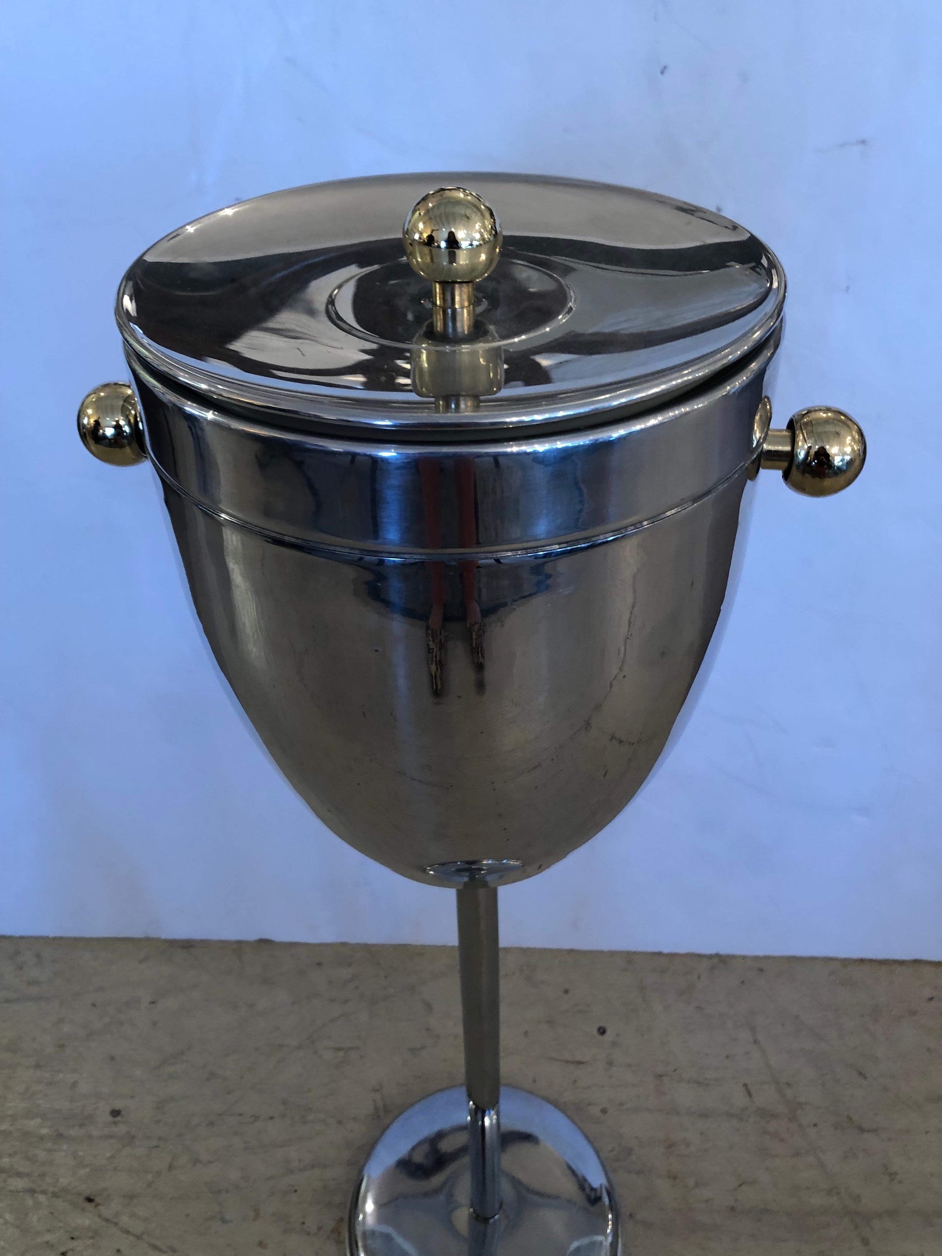 A glamorous and functional midcentury modern chrome champagne ice bucket on a stand having brass shaped ball handles and finial on top.