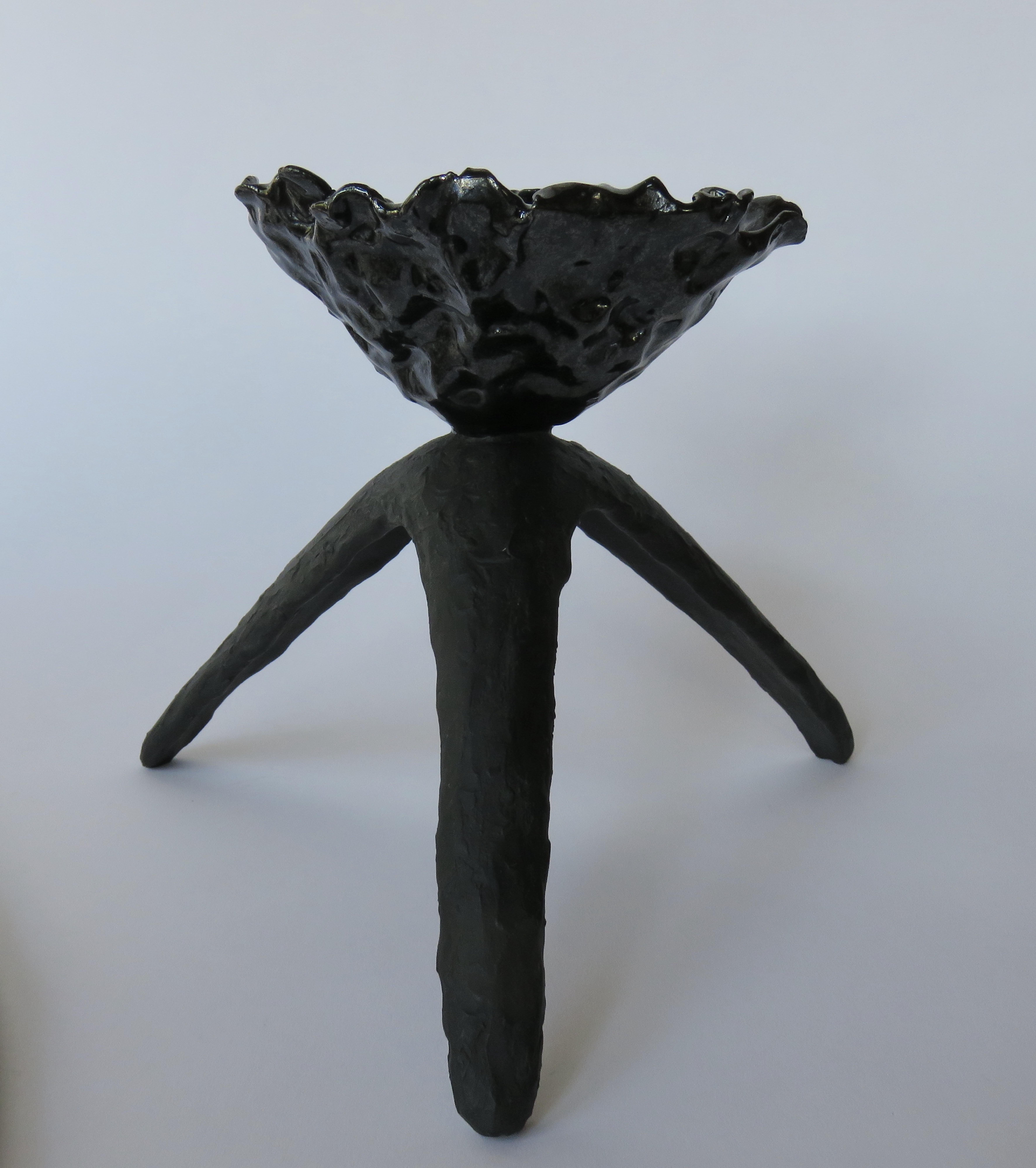 A freeform ceramic chalice with glossy black glaze on top and matte black tripod legs. The top 'cup' was pinched and crimped into this abstract form, then coated with a dramatic black glaze that shows all the highlights and movement in the piece. It