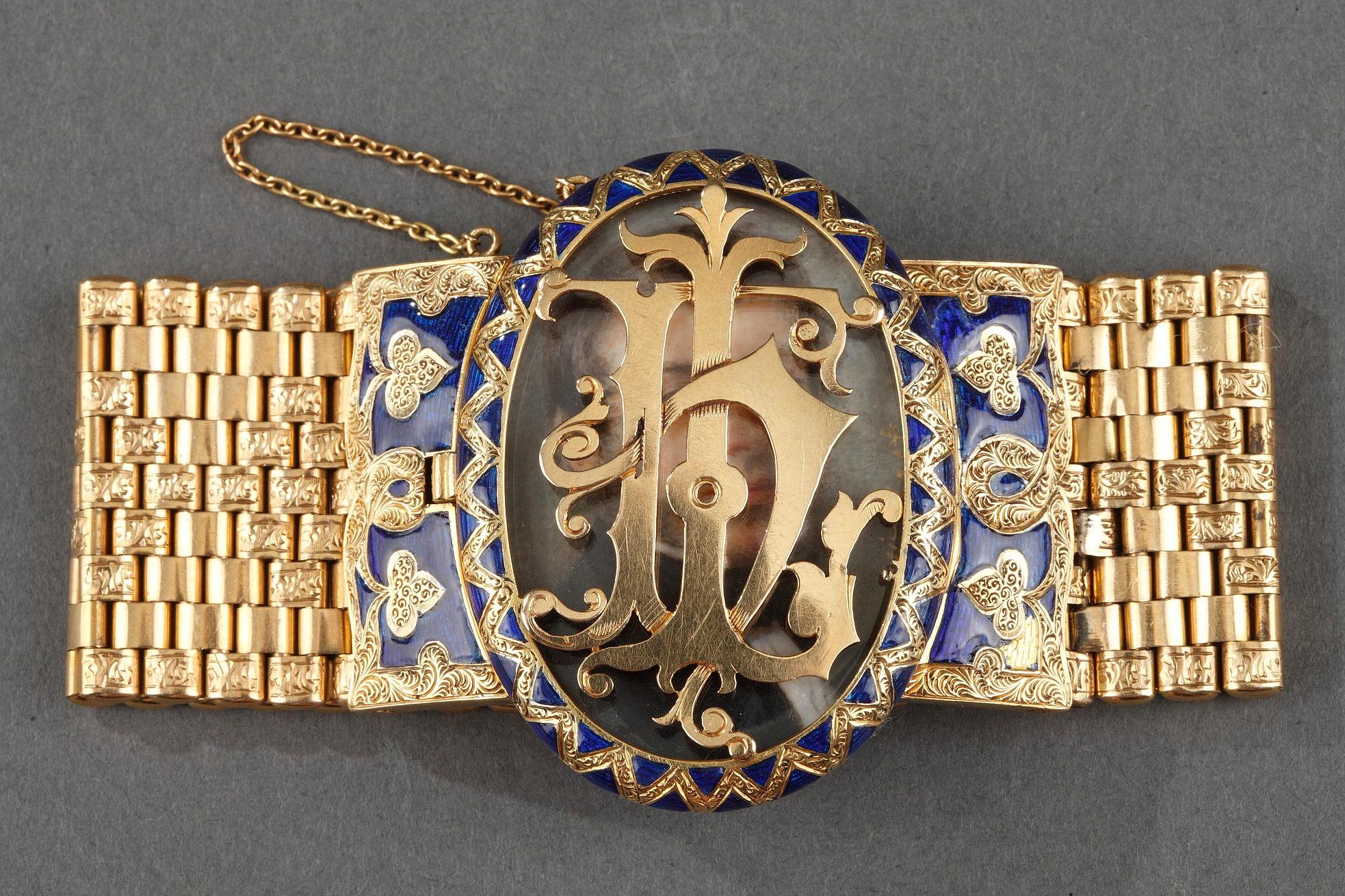 The strap of woven gold with stylized patterns arranged alternately and decorated at the ends with oval medallion surrounding with blue enameled gold and a hinged openwork cover to protect a miniature. The cover features a monogram. The miniature