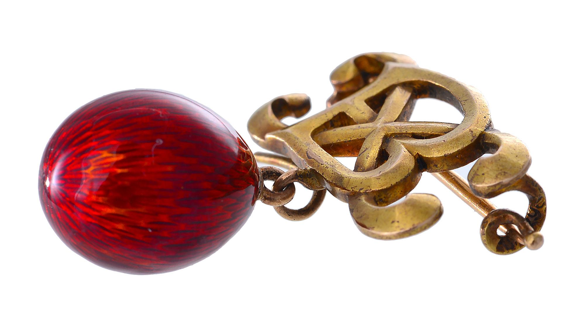 A brooch of a gold cyrillic letters depicting the message 'Christ Is Risen' suspending a small red Cloisonné enamel egg. The brooch has a makers mark attributed to the Faberge jeweller Alfred Thielmann. The brooch is marked on the clasp A.T and the