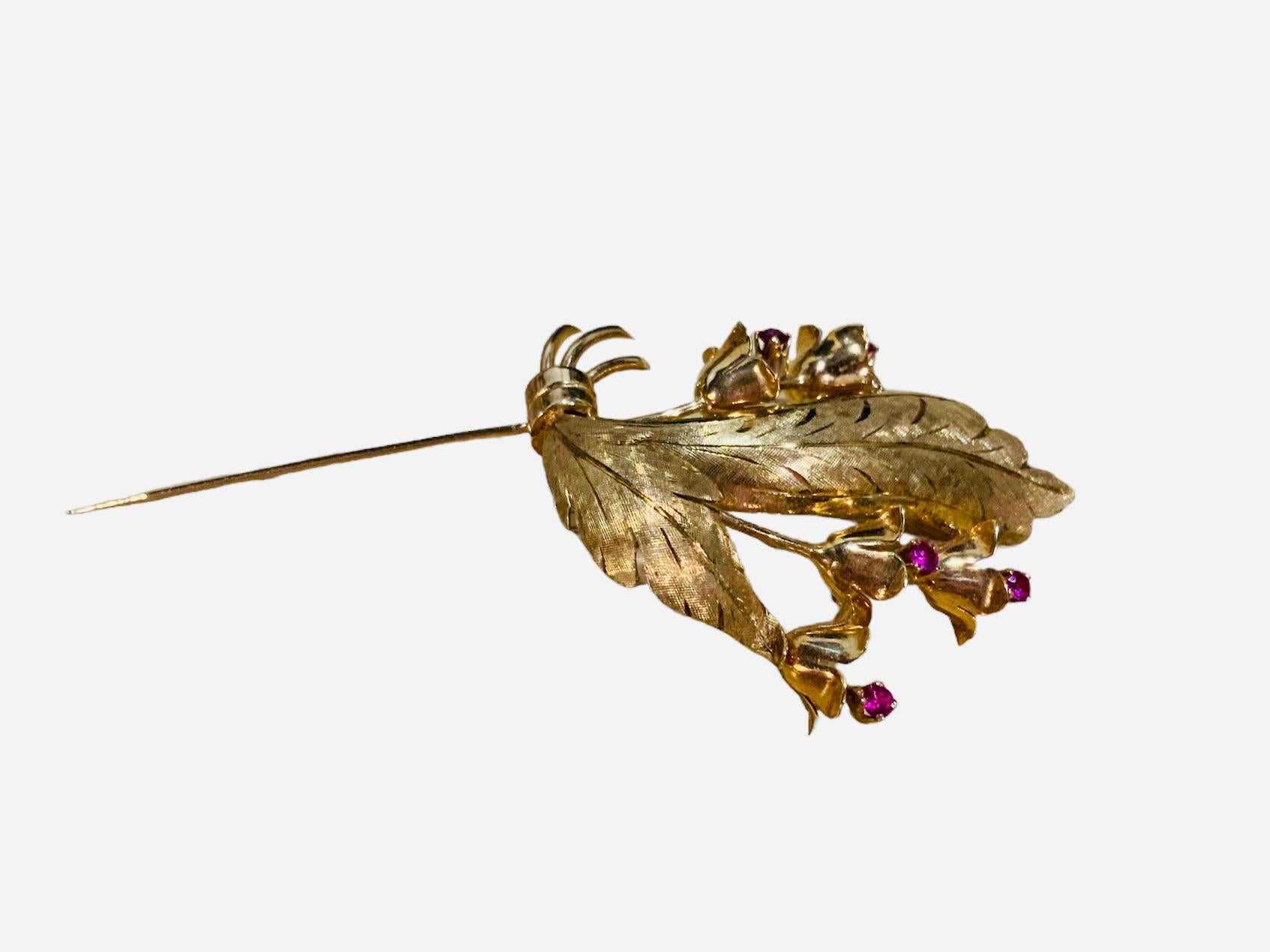 This is a yellow gold and rubies brooch. It depicts a bouquet of three branches of Lilies of The Valley with five flowers and two leaves hold by a ribbon. Each flower is embellished by a round cut ruby in prong setting. The brooch has in the back a