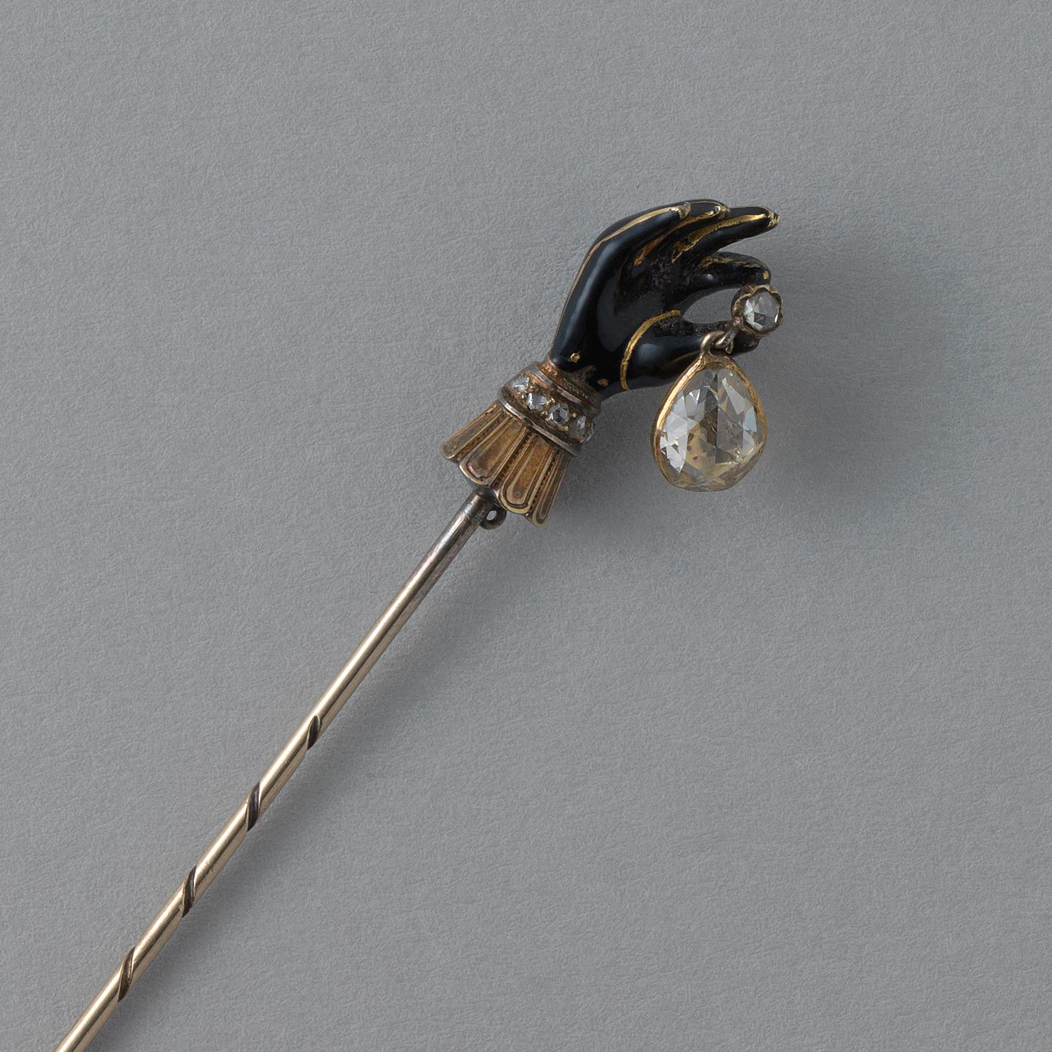 A rare neo-Renaissance stickpin or ‘ear iron’ featuring a gloved hand decorated with black enamel with gold seams. The thumb and index finger hold a small rose-cut diamond, from which dangles a pear shaped rose-cut diamond. At the top of the gold