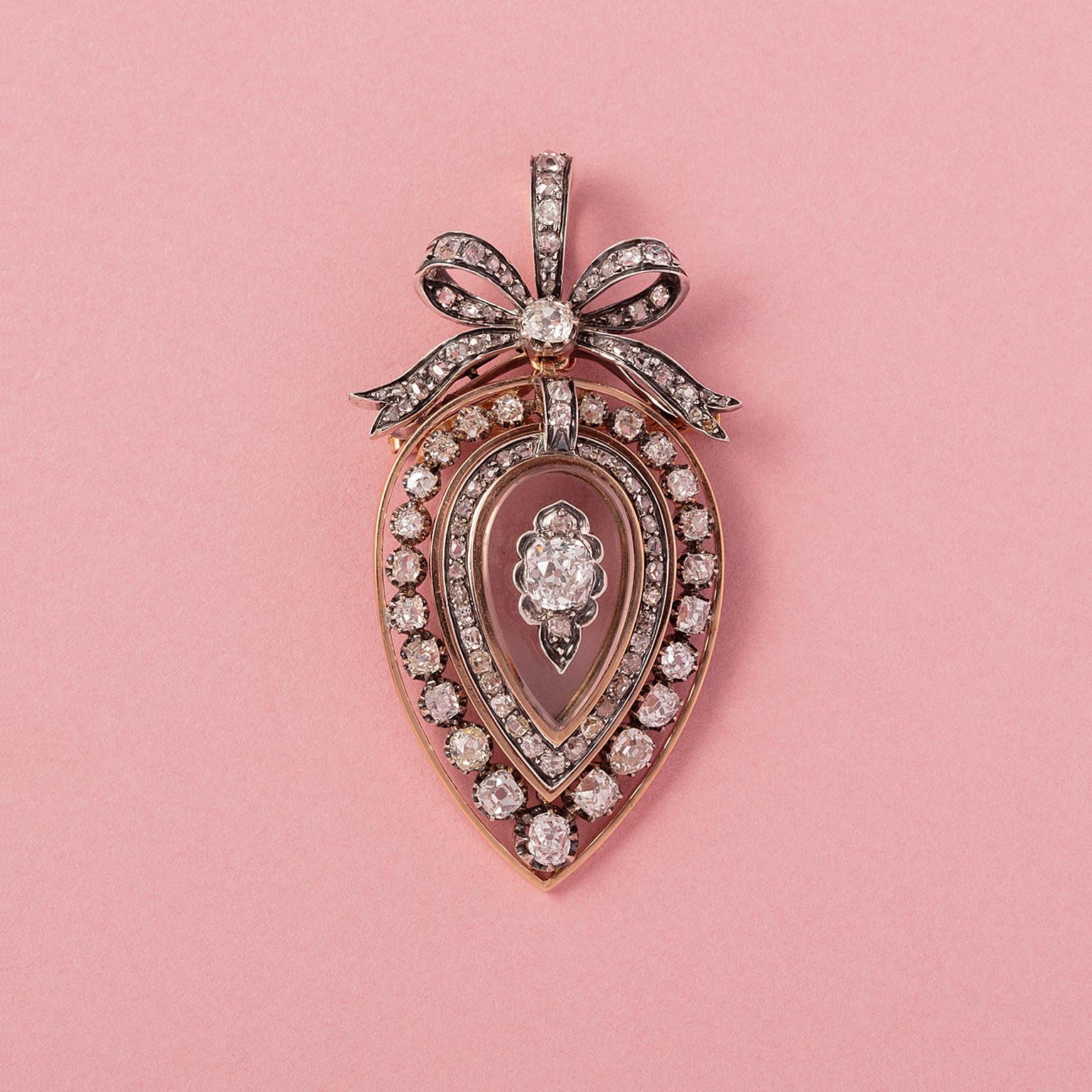 An 18 carat gold and silver locket pendant (or brooch with brooch fitting) in a stylized heart or drop shape with an elegant bow on top. In the middle a drop shaped rock crystal incrusted with a cushion cut diamond (app. 0.9 carat I-J SI 2) and two