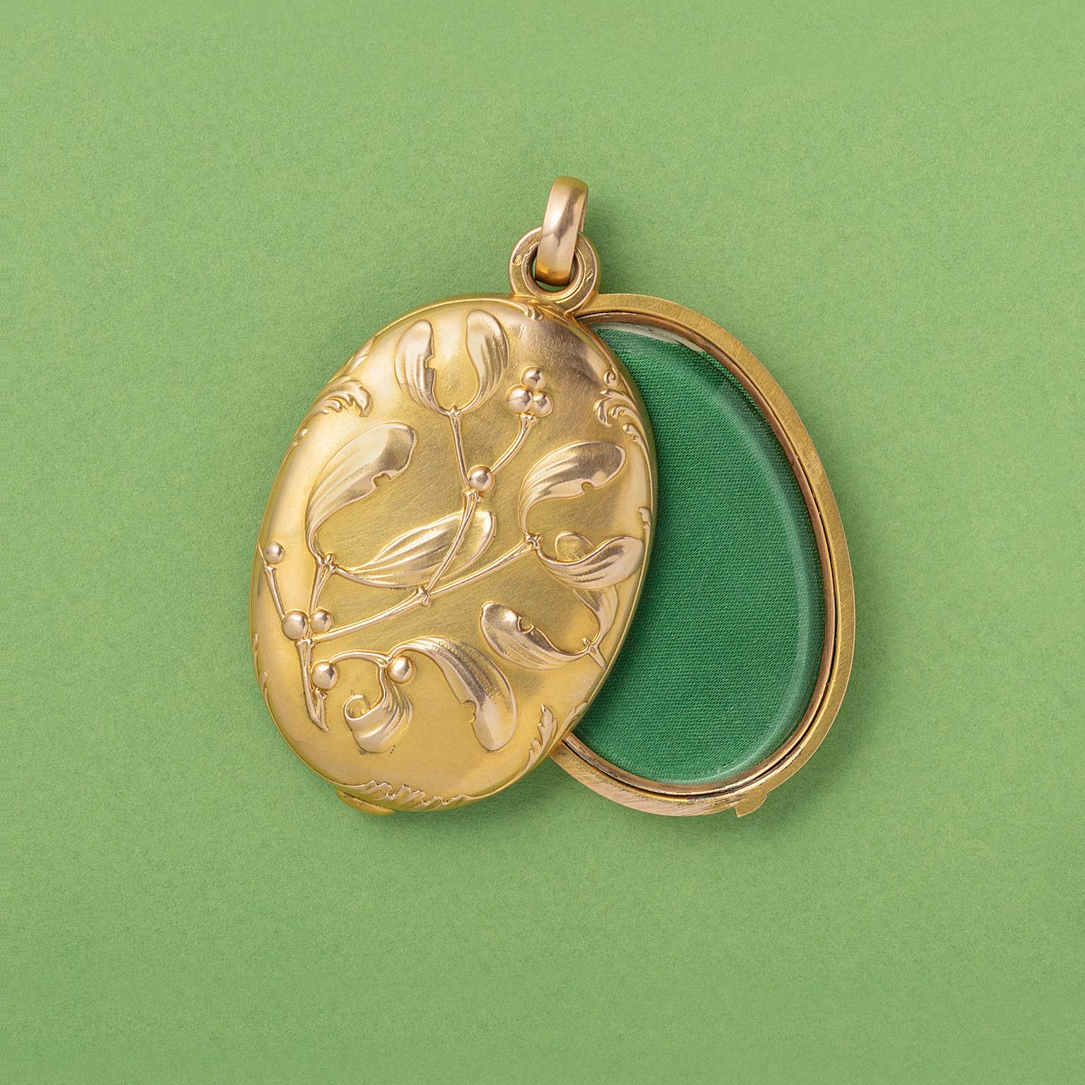 A large, oval, 18 carat yellow gold locket with gold missletoe on front and back 'en relief'. The locket opens sideways and on the inside a mirror on one side and a glassed compartment on the other. Mistletoe symbolises constancy and stands for a