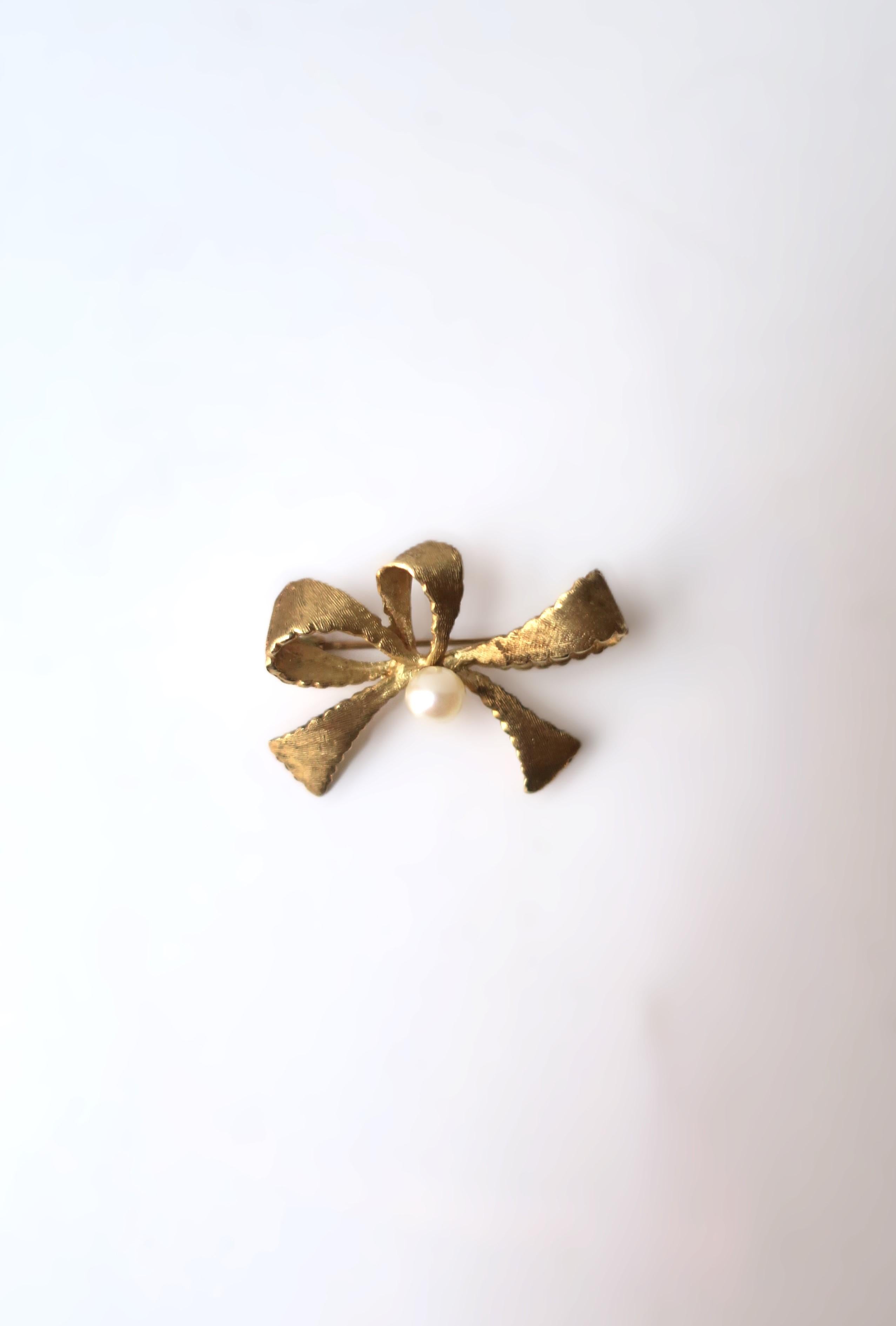 A gold bow and white cultured pearl pin brooch in 14 karat yellow gold, in the style of Tiffany & Co., New York, circa 1960s. A beautiful textured 14-karat yellow gold bow with white cultured pearl at center. A great piece for a sweater, coat,