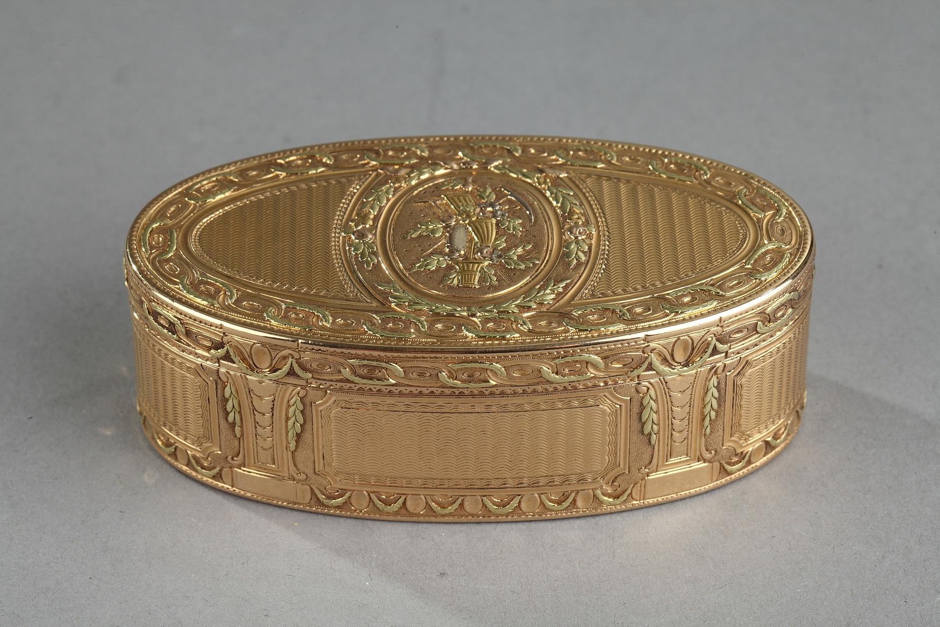 Three-tones gold oval snuff box. The hinged lid is decorated with a country trophy medallion. The medallion is framed by a garland of green gold foliage and roses. The medallion is detached on a guilloché. The borders of the lid and the box are