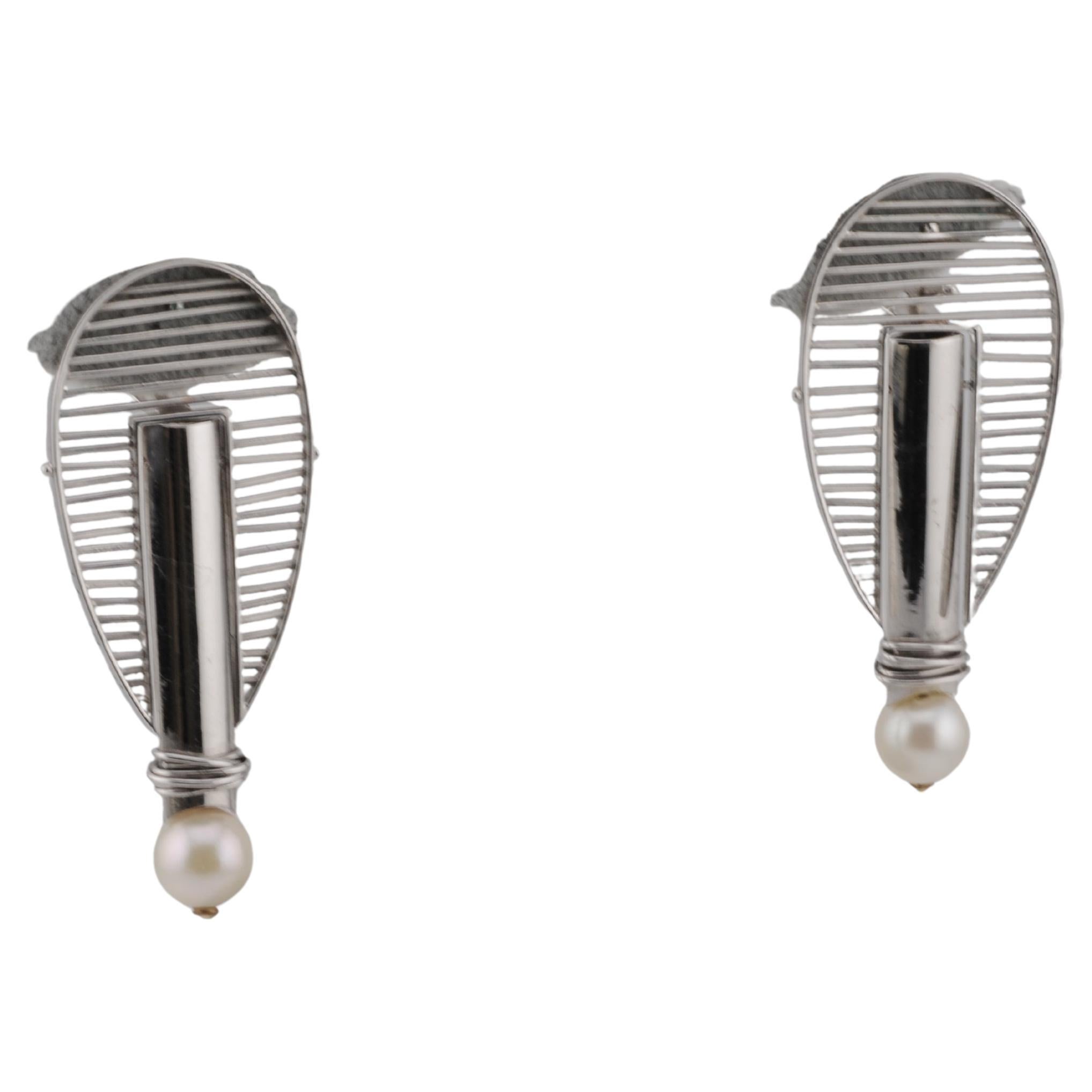 Orecchini pendenti in oro bianco 18 carati, realizzati a mano.

Il design segue i principi del Bauhaus: pulito, geometrico e funzionale. Ogni orecchino presenta una perla fine. Gli elementi mobili aggiungono un leggero movimento quando vengono