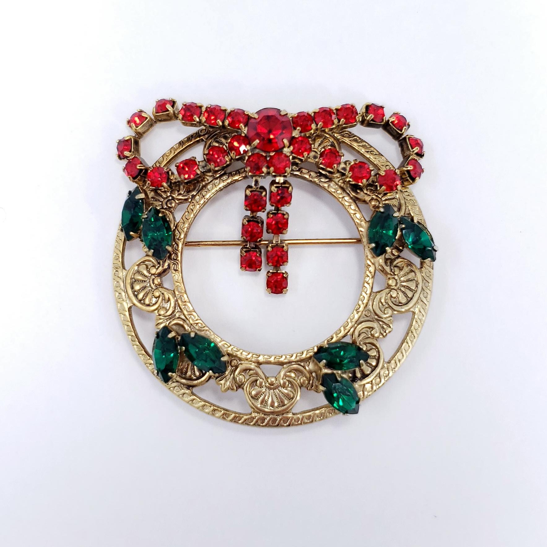 A festive pin brooch, featuring a gold-plated wreath decorated with a red-crystal bow and green crystal accents.

Hallmarks: n/a