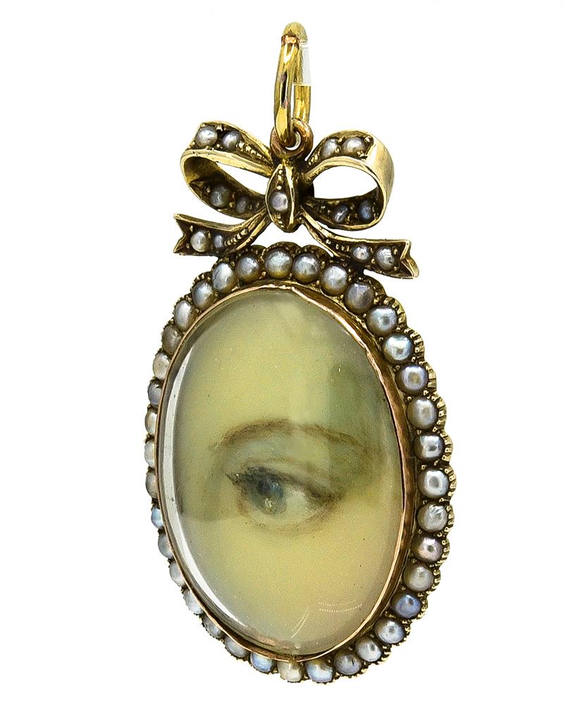 A romantic example of the rare Georgian Lover's Eye jewelry:  a pendant with a fine hand-painted eye.  Set in an oval mounting
surrounded by seed pearls, suspended from a bow.  9K yellow gold.  2