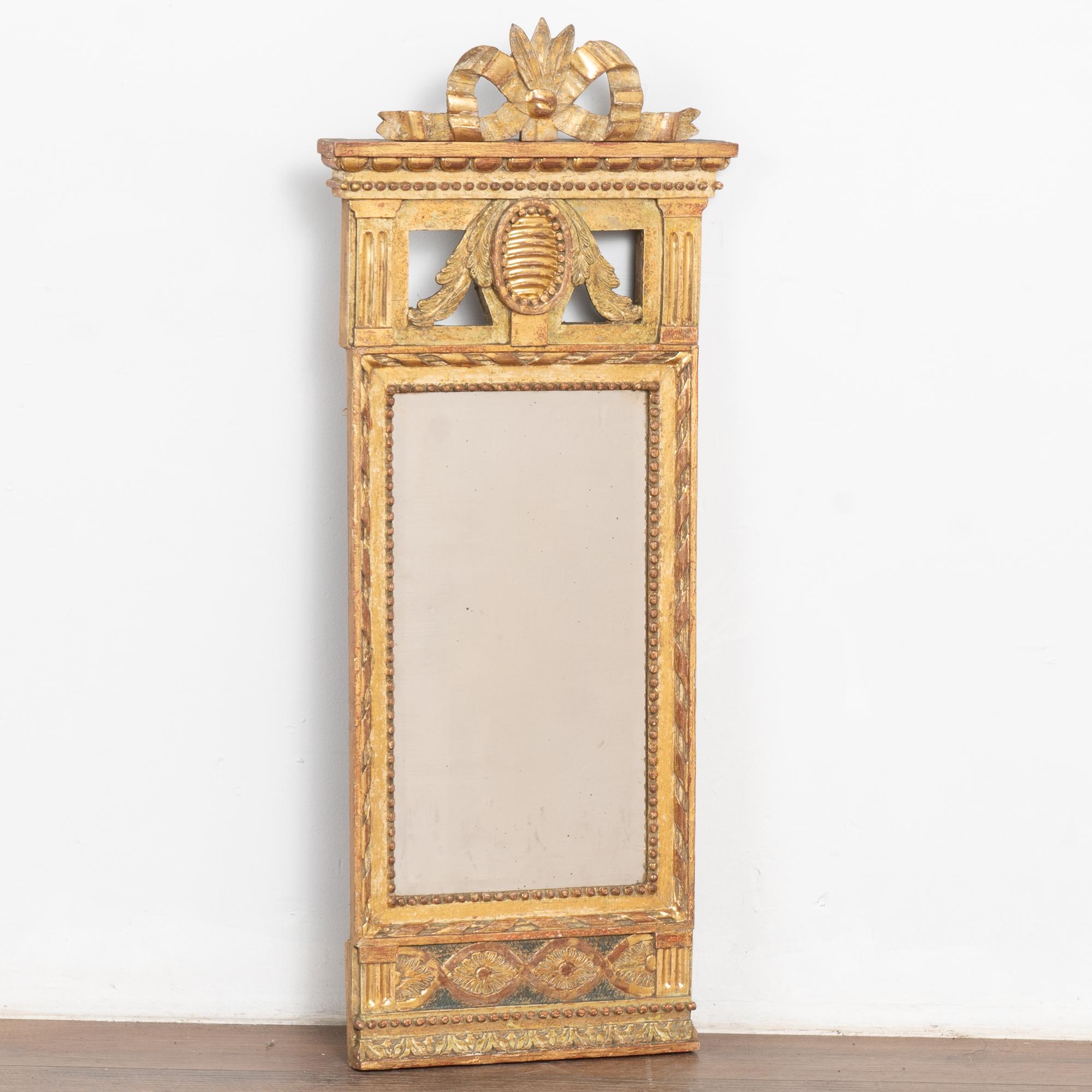 Joli miroir trumeau étroit avec un motif de ruban peint et sculpté couronnant la partie supérieure, une moulure de denticules, une garniture de perles et plus encore.
De magnifiques nuances d'or, de vert pâle et de rouge bruni sont parfaitement