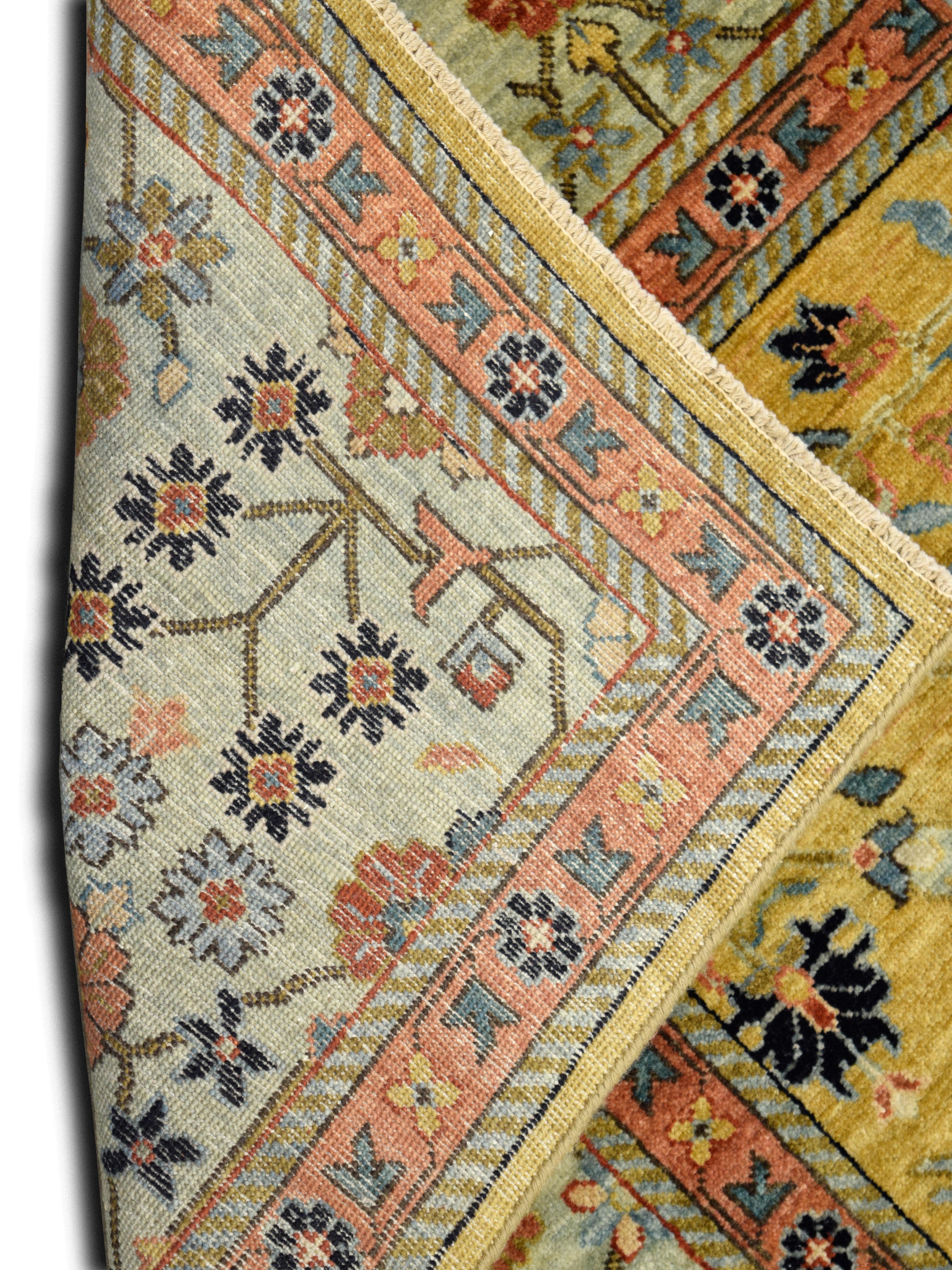 This gold handknotted rug with a blue border, pink, green, and floral motifs, measuring 6' x 9', captures the beauty of timeless design with a vibrant and intricate composition. The glowing golden field is adorned with delicate floral patterns in