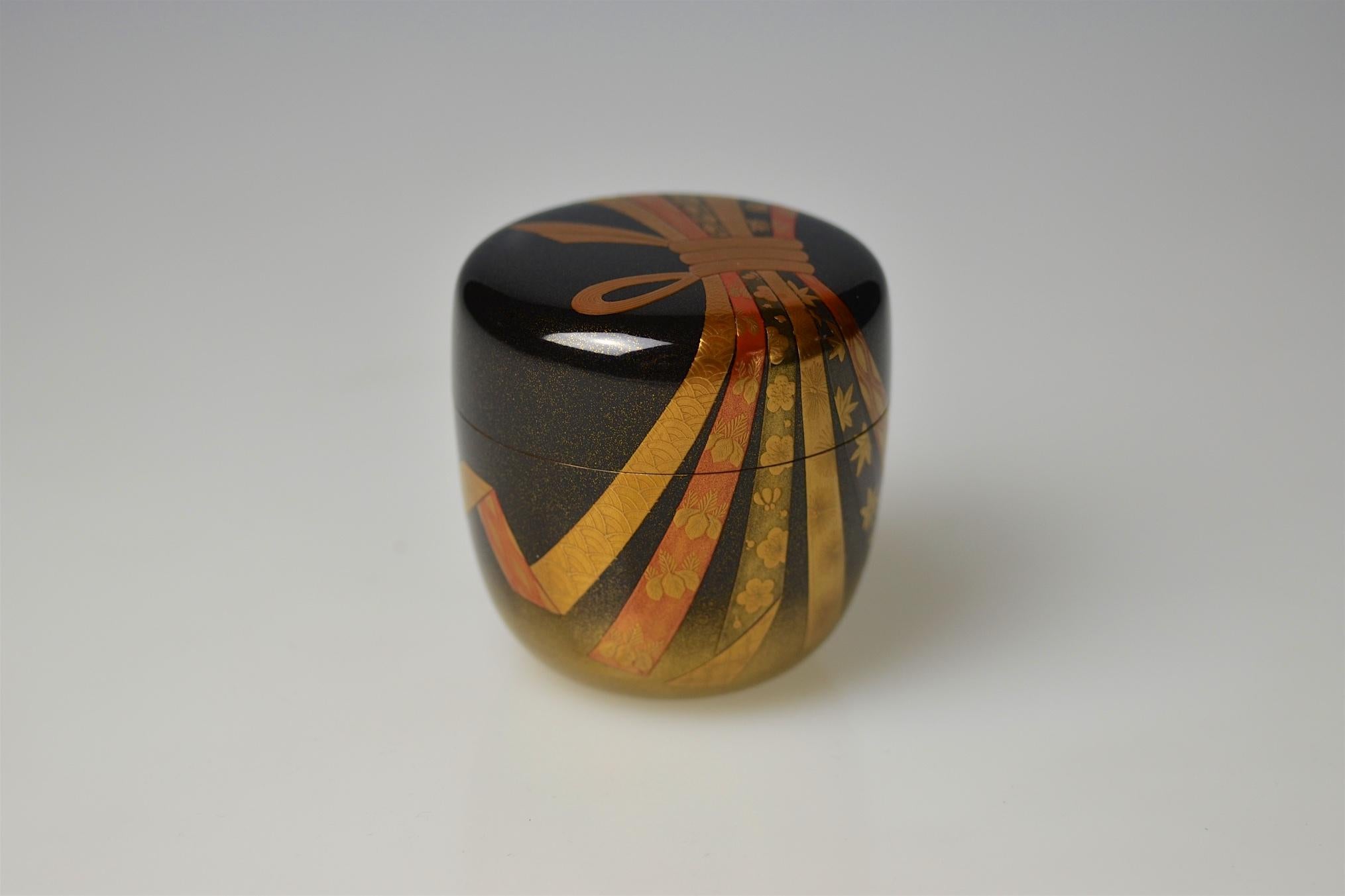 A large tea caddy (ô-natsume) made by Ichigo Itcho (1898-1991) with decor of festive ribbons that are hold together by a golden knot (noshi). The wood body is finished in polished black lacquer (roiro) and sprinkled with gold powder in the lower