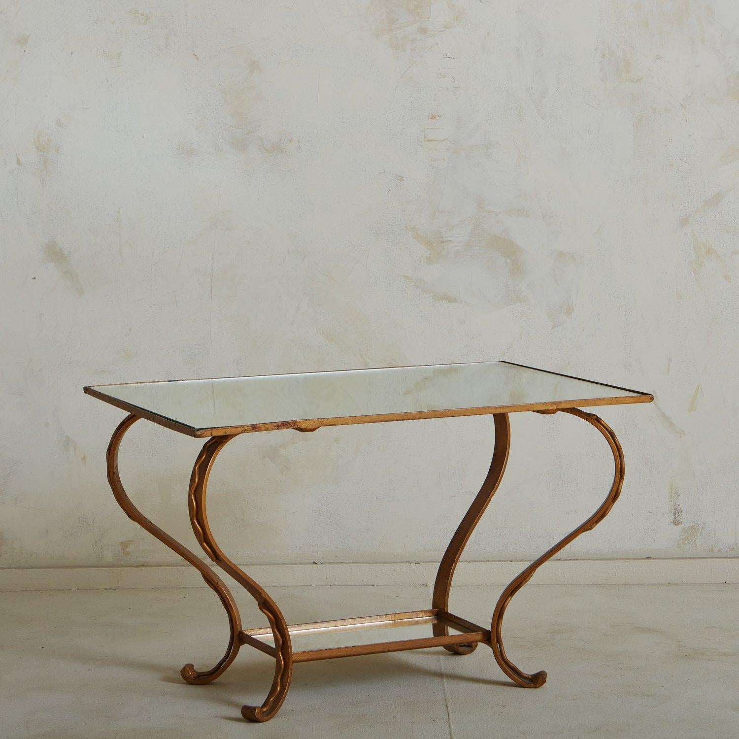A 1950s French coffee table featuring a metal base with a beautiful hand applied gold leaf finish. This table has curved legs, which have an overlaid metal squiggle design. It has an inset rectangular mirrored tabletop and a lower mirrored tier