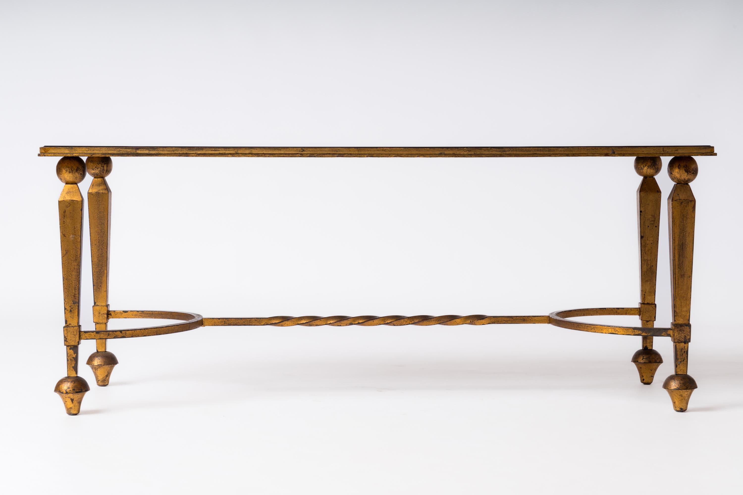 Table basse néo-classique graphique attribuée à la Maison Ramsay présentant une base architecturale en fer recouverte de feuilles d'or et un plateau en verre opalin noir. 
Pertes de feuilles d'or et patine sur toute la base, comme le montrent les