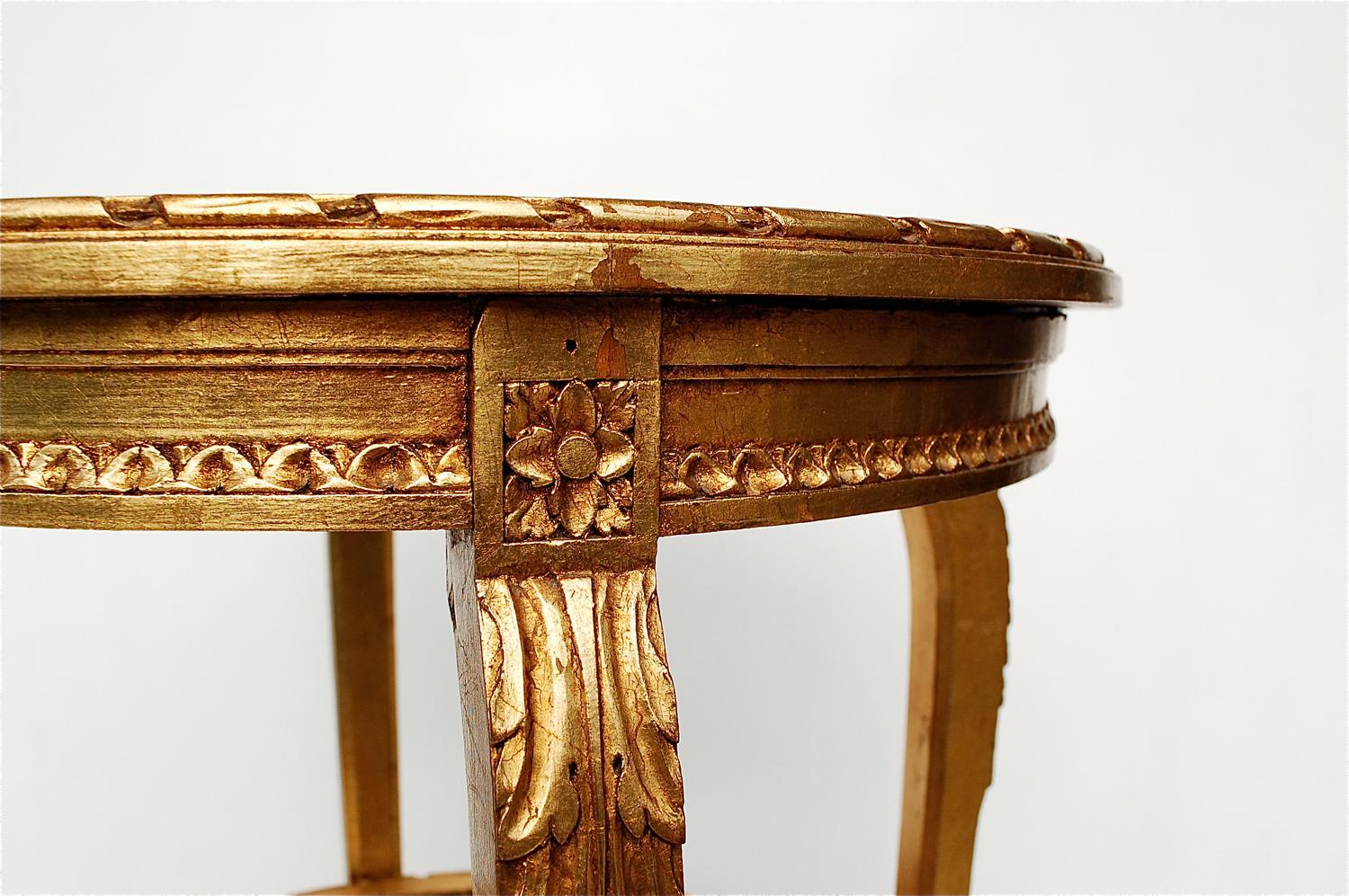 Louis XIV, French Empire style side table on tall, slightly curved legs. It has a carved wooden frame with leaf motifs at the top of each post. The wooden frame has been painted gold, giving it a warm, rich glow. The tabletop has a circular mirrored