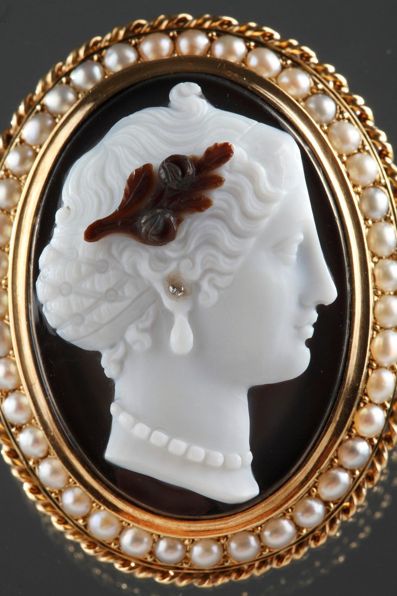 19th century oval agate brooch. It depicts the profile of a woman wearing a necklace and diamond earrings, set within a golden frame and accented with a ring of pearls. The sculpting of this brooch is very finely done. 

French mark “tête d’aigle”