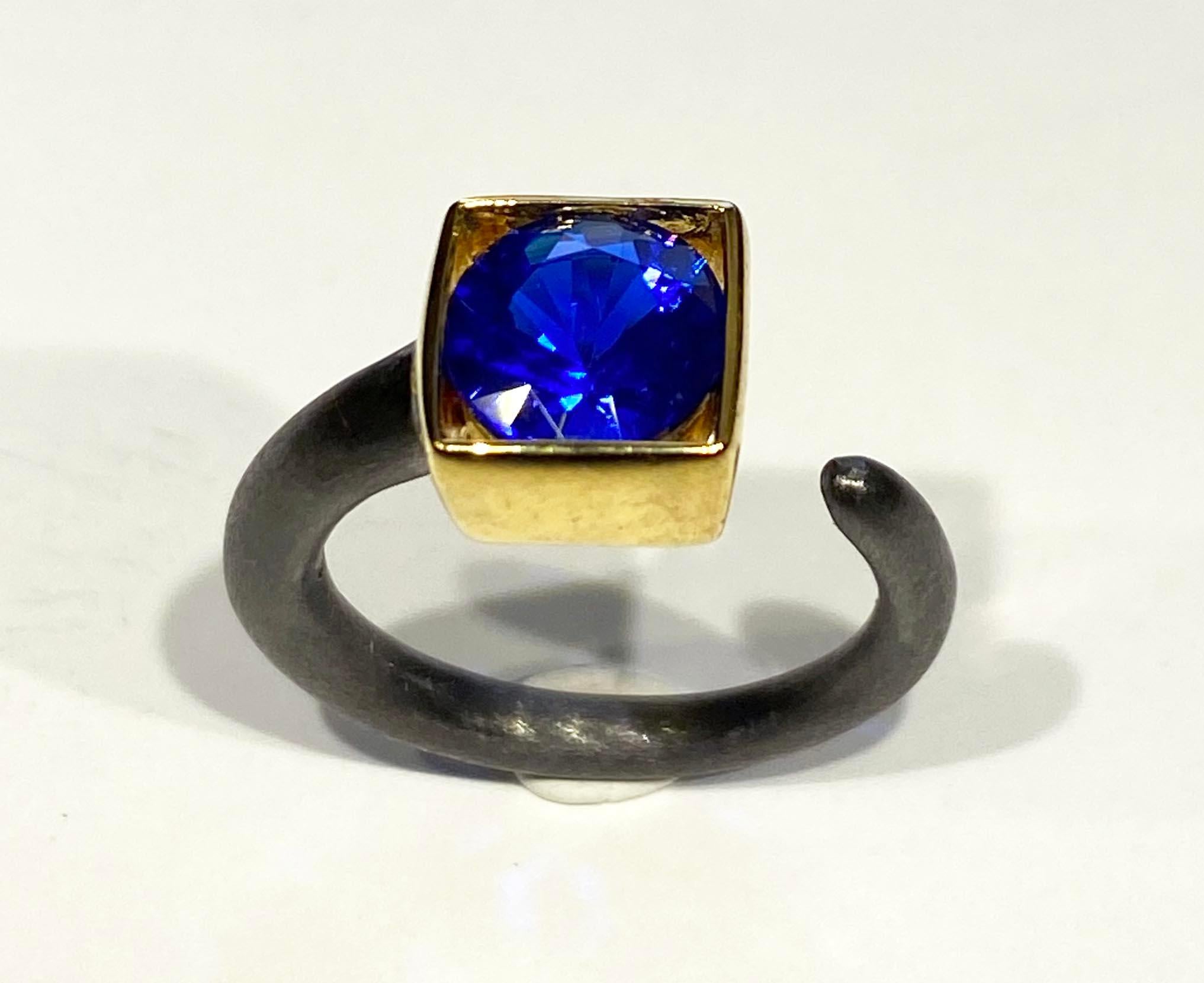 Gold Plated Blackened Silver Ring with YAG Blue Garnet. 
This Curl style Blackened Silver and Gold Plated Ring features a Box Set Vivid Blue Yittrium Aluminum Garnet of 1.6 Carats 8MM Round. The ring is Sized at 6.5 US.

Originally from San Diego,