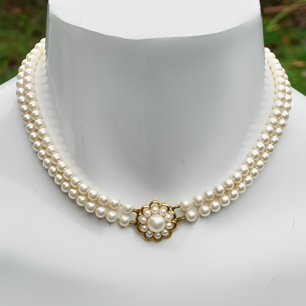 Splendido collare placcato oro con due fili di perle finte color crema e una catena di estensione. Il ciondolo ha un grazioso disegno floreale.

Misura circa 33 cm di lunghezza con un'estensione di 7 cm di lunghezza. La larghezza è di 1 cm.

Questo