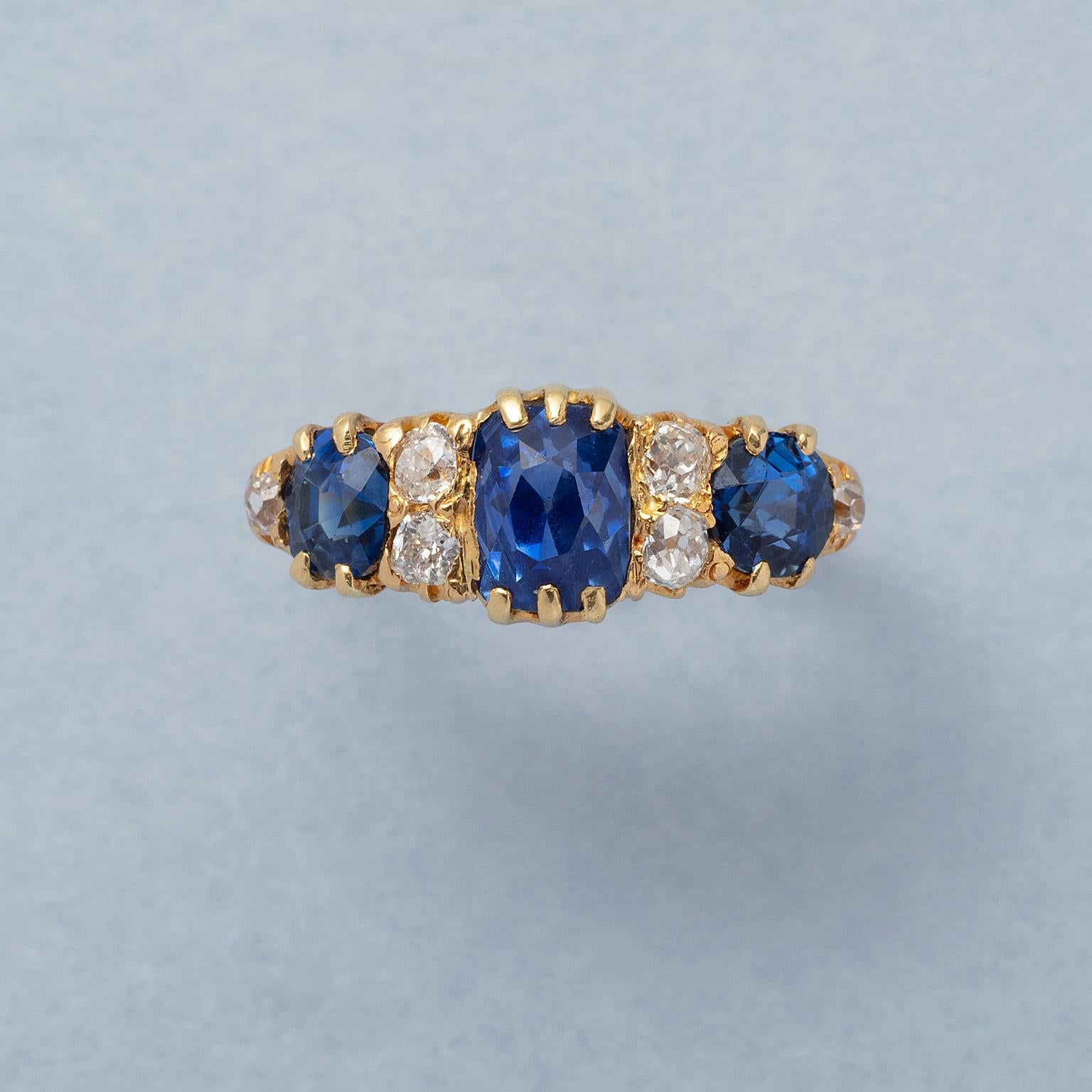 Ein Ring aus 18 Karat Gelbgold mit drei königsblauen, natürlichen, unerhitzten Saphiren mit jeweils zwei Diamanten im Kissenschliff dazwischen; insgesamt 6 Diamanten im Kissenschliff (ca. 0,42 ct). In der Mitte ein Saphir im Kissenschliff mit einem