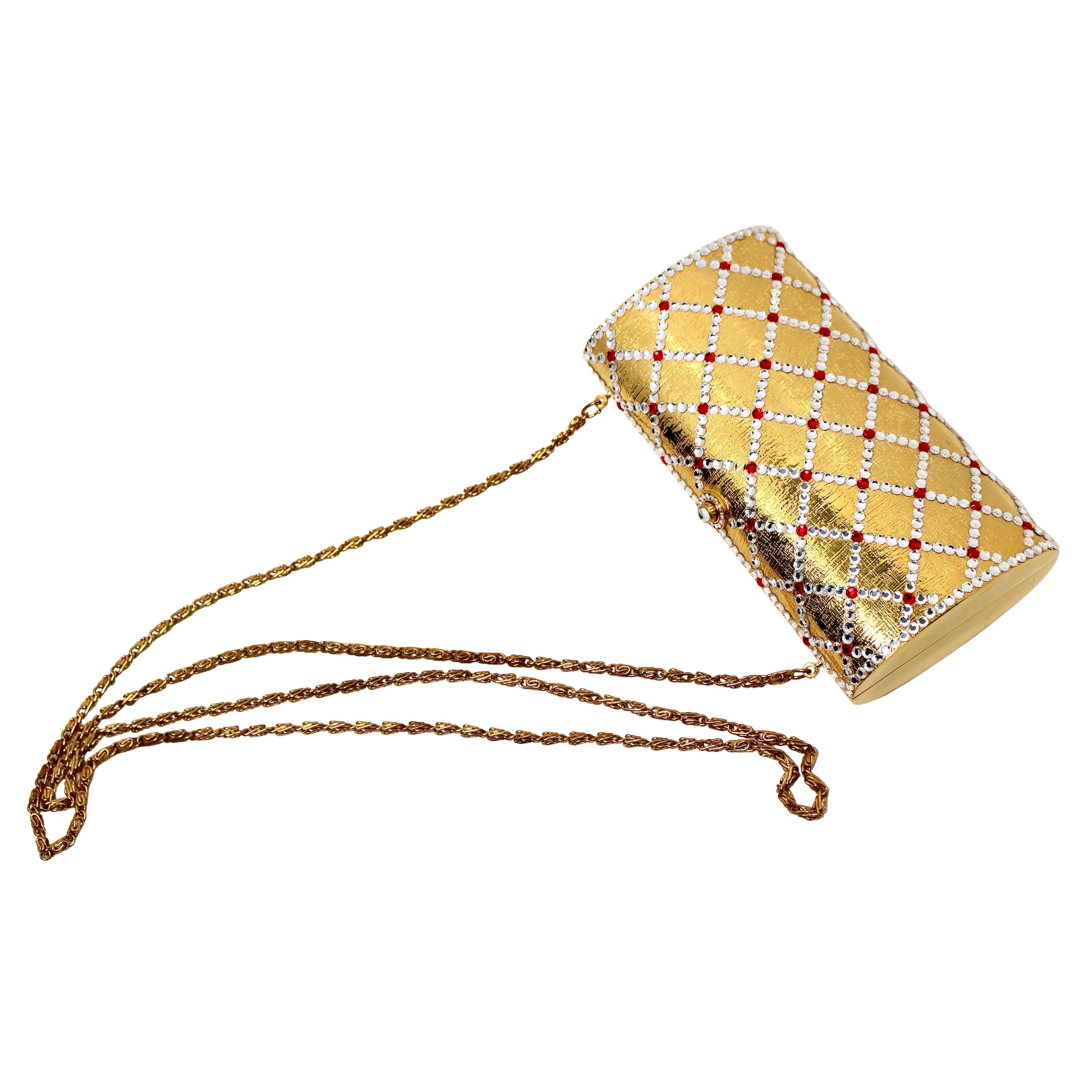 Cette superbe pochette vintage des années 1960 présente un luxueux corps en métal doré orné d'une multitude de strass rouges et transparents scintillants, disposés selon un motif matelassé complexe. Les ornements lui confèrent un aspect glamour et