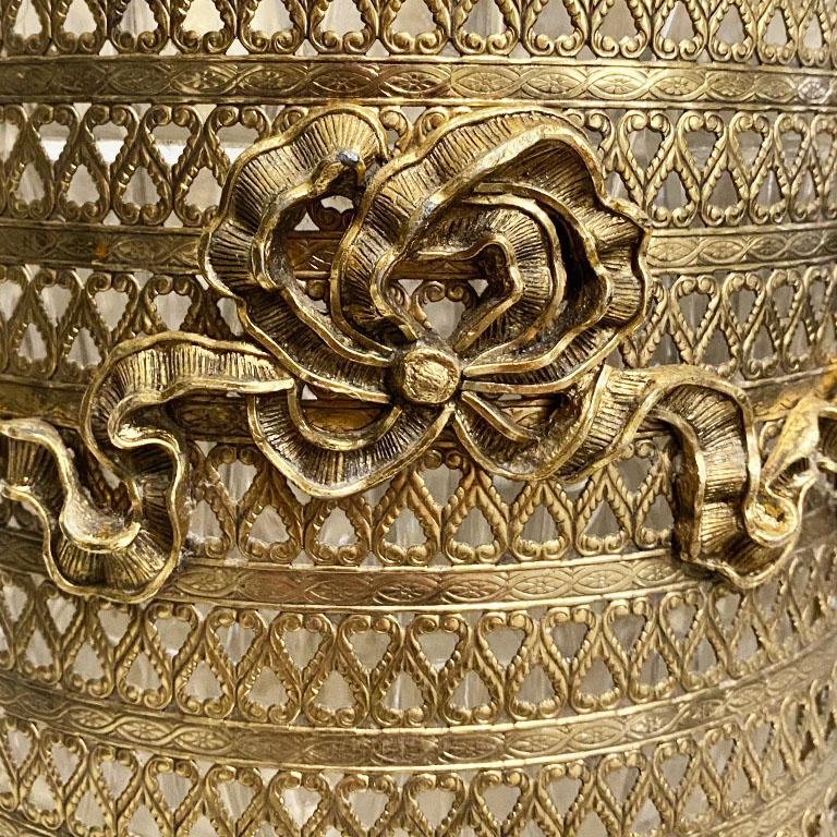 A gold pierced metal wastebasket with trompe l'oeil ribbon detail and claw feet. Around the sides, the bin features pierced heart-shaped details. The center of the bin showcases a trompe l'oeil ribbon also in gold. The bottom has claw feet. The bin